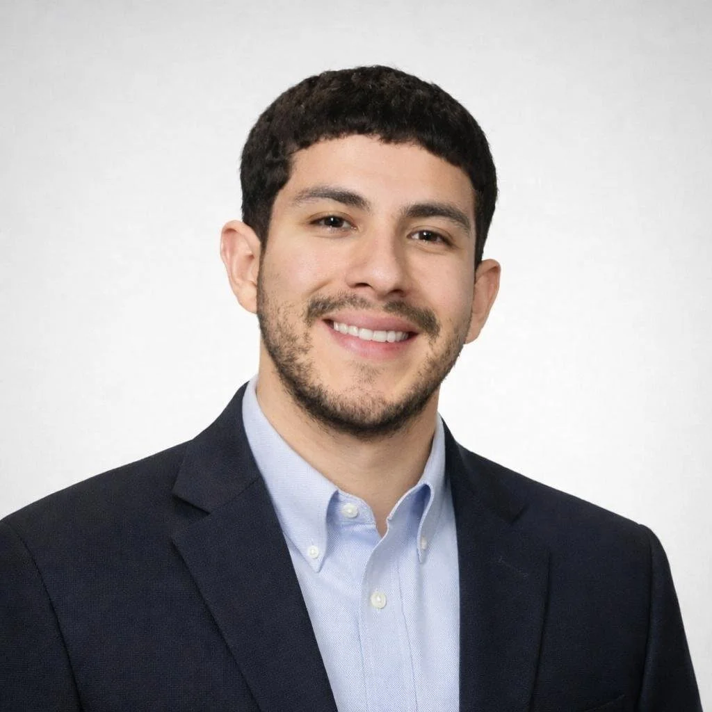 Portrait of a young man with dark hair, beard, and mustache wearing a suit jacket over a light blue collared shirt, smiling against a plain background.