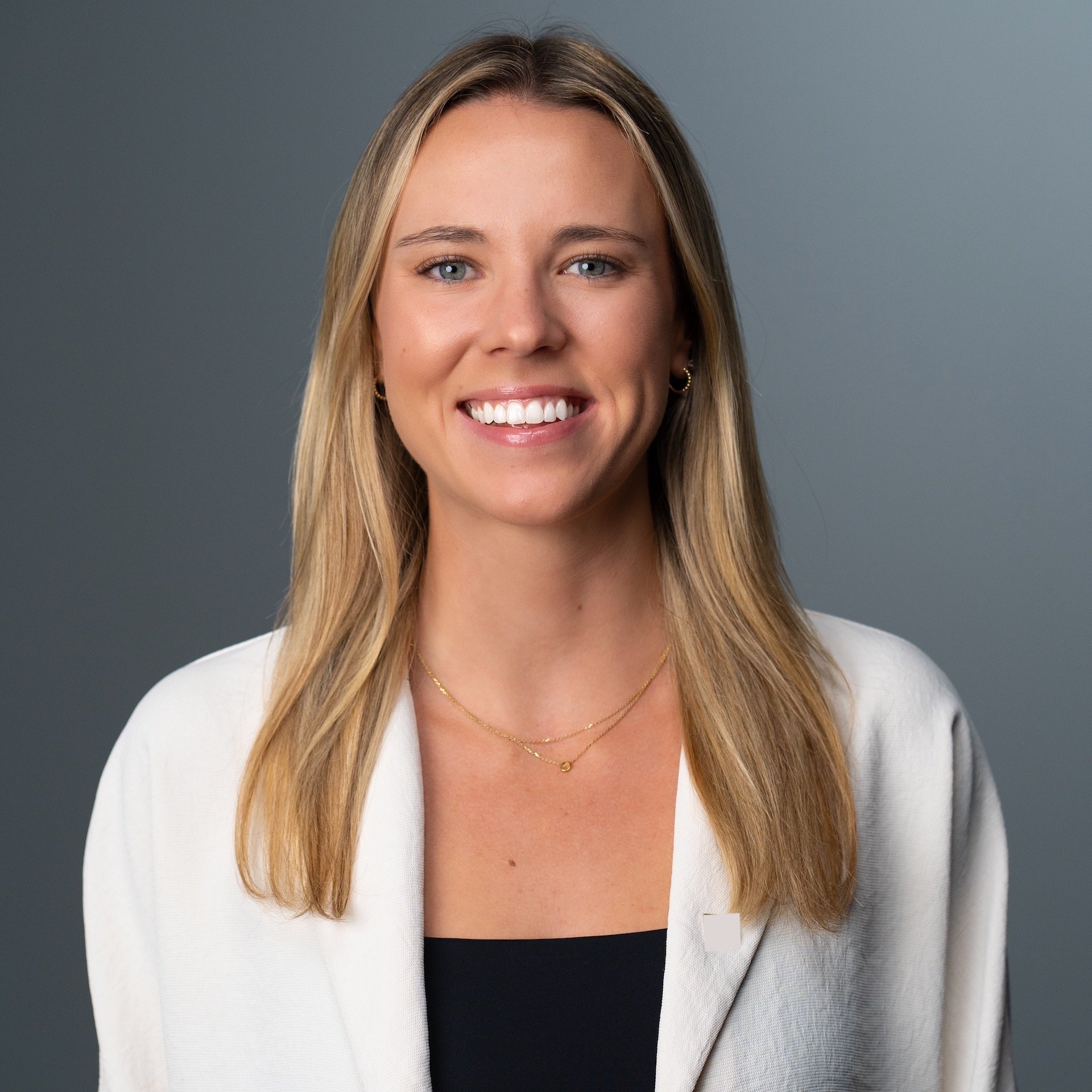 Portrait of a young woman with long blonde hair, blue eyes, smiling, wearing a white blazer and layered jewelry, against a gray background.
