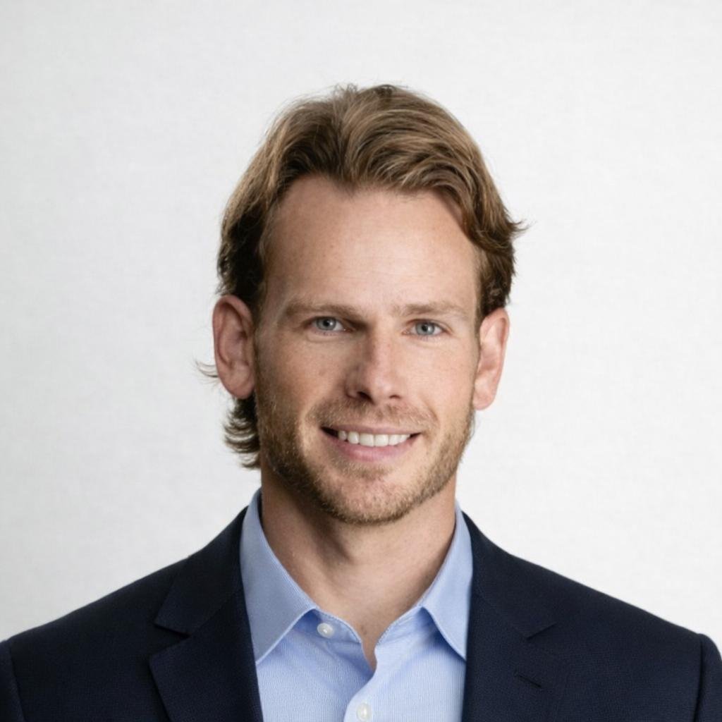 Portrait of a man with light brown hair, blue eyes, and a beard, wearing a light blue shirt and a dark blazer, smiling against a plain white background.