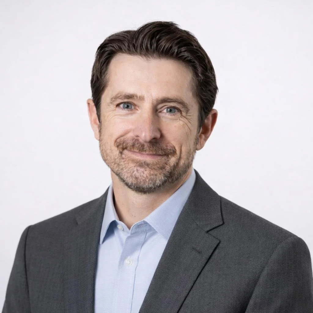 Headshot of a man with dark hair, blue eyes, and a beard, wearing a gray suit and light blue shirt, smiling against a plain white background.