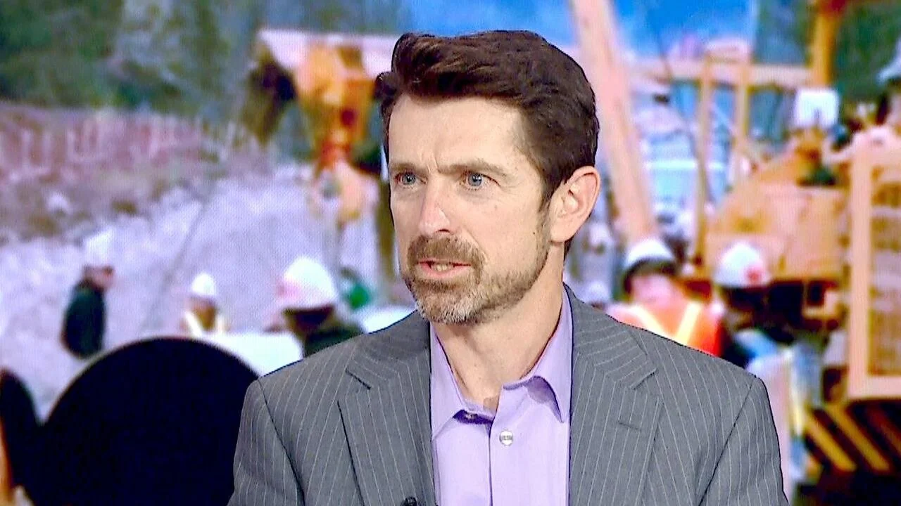 Man with brown hair and beard wearing a gray pinstripe suit and lavender shirt in front of a construction site with workers in hard hats and construction equipment.