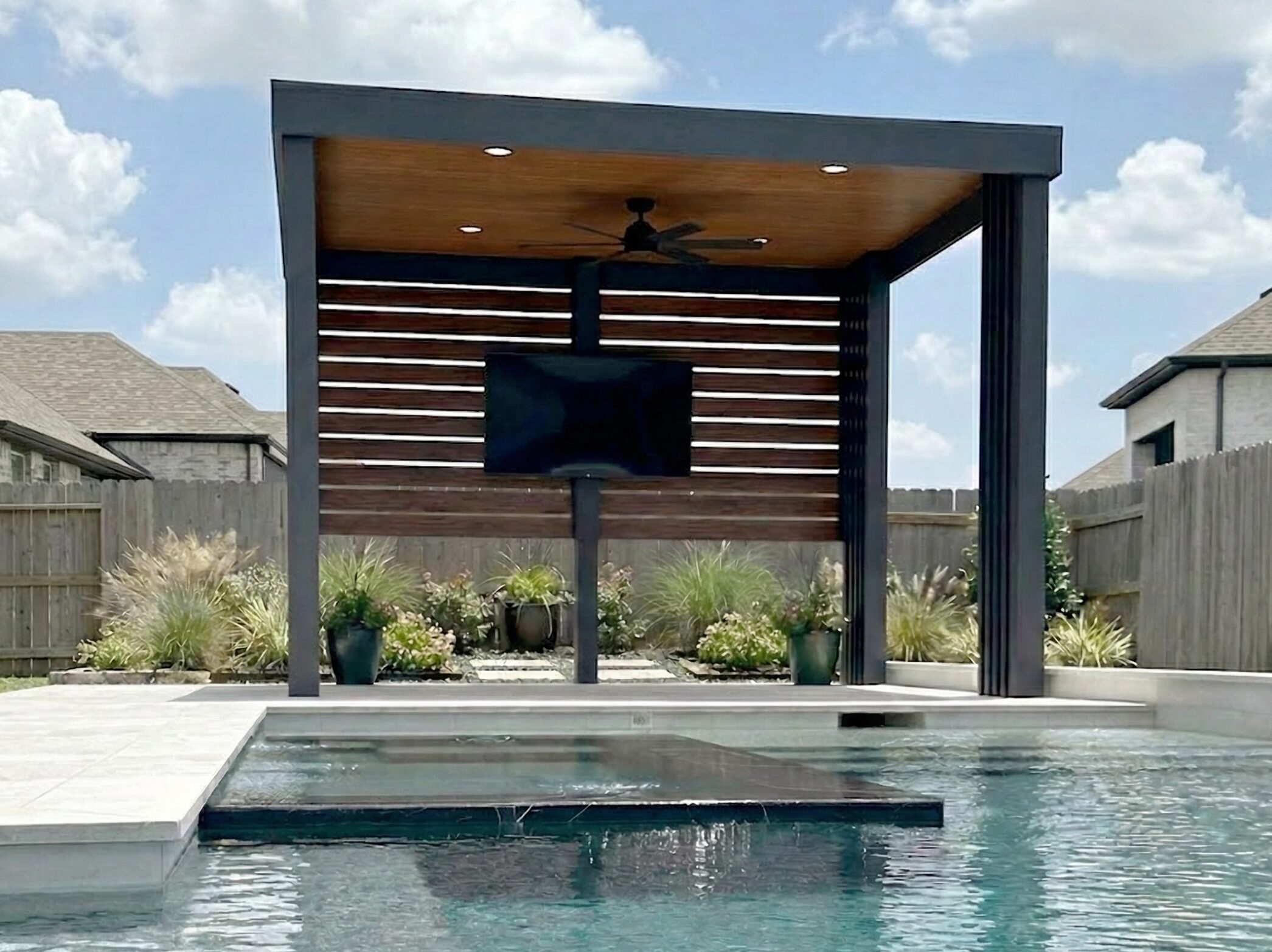 A backyard with a modern structure featuring a wooden ceiling, black finials, a ceiling fan, and built-in lighting. There is a wooden privacy screen, a television mounted on the structure, and a pool in the foreground with a well-maintained garden and neighboring houses visible in the background.