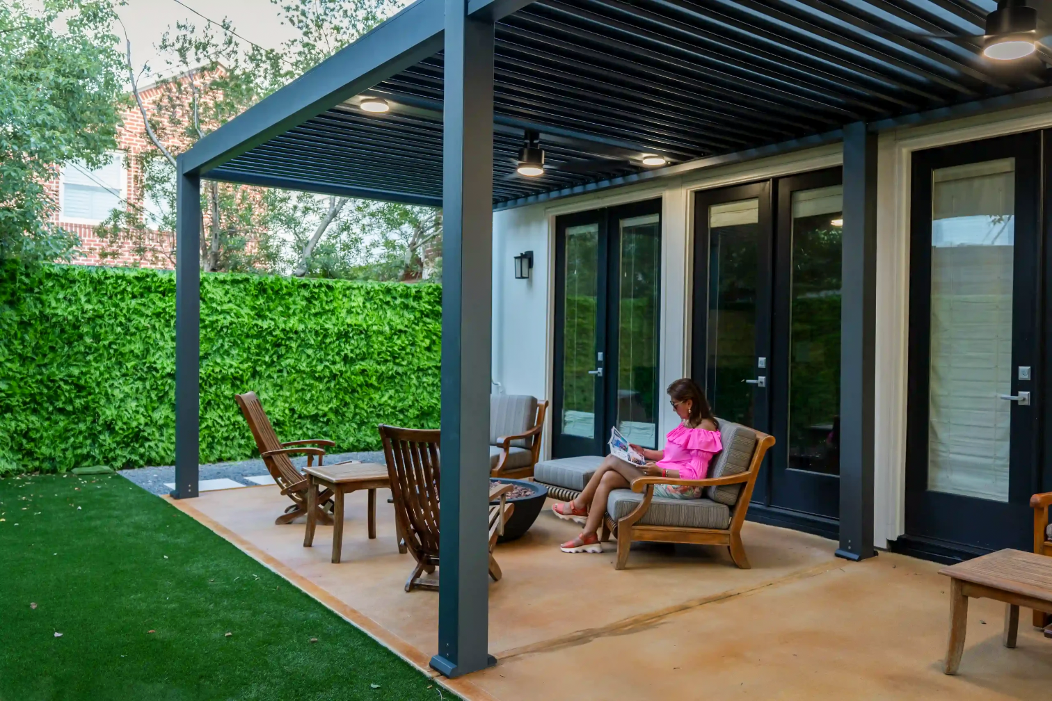 A woman in a pink dress sitting on an outdoor patio chair, reading a magazine, under a modern black metal pergola attached to a house with sliding glass doors, surrounded by a green hedge and lawn.