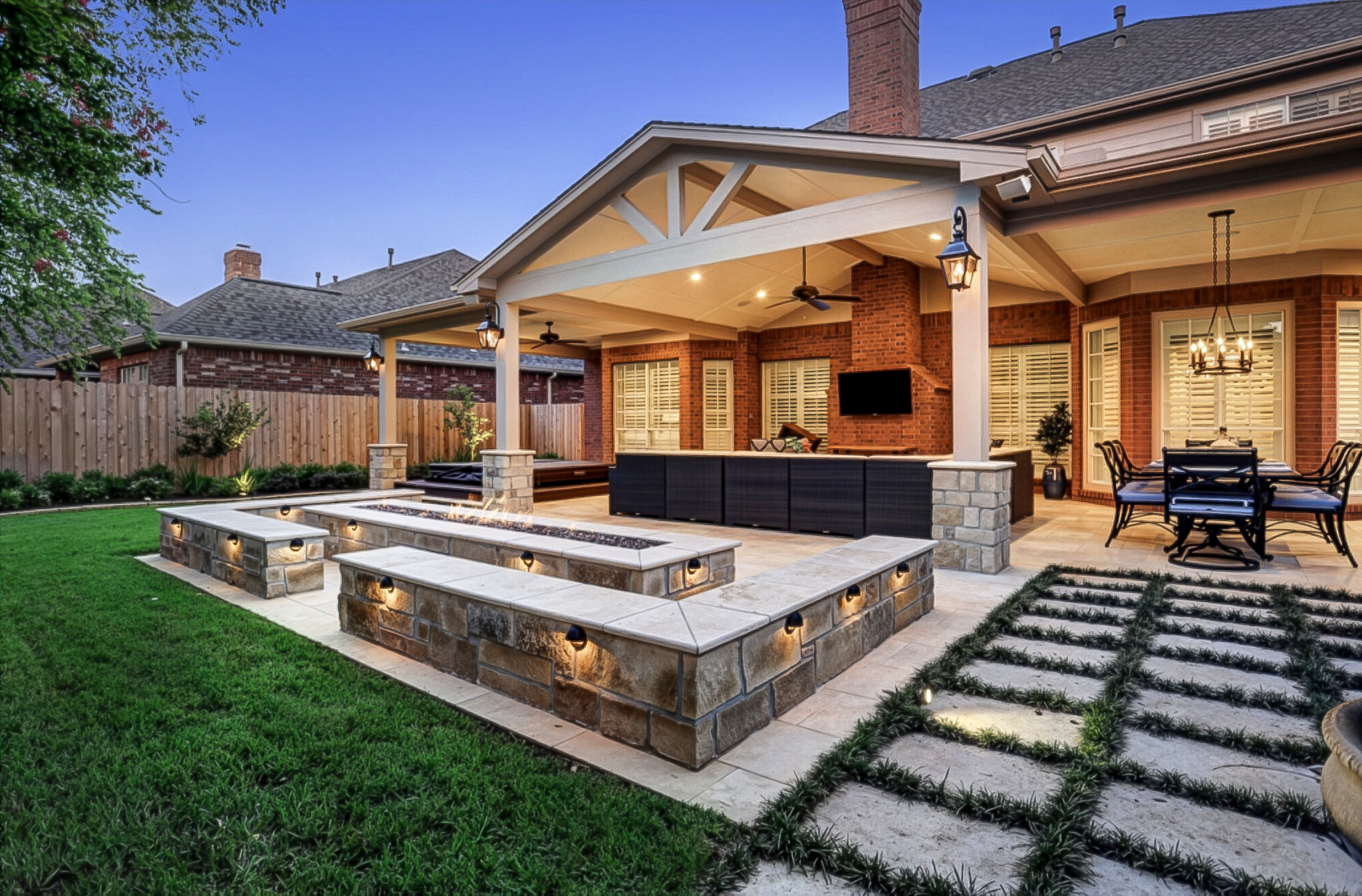 Backyard patio with brick and stone features, outdoor furniture, hanging lights, and a fence, during dusk.