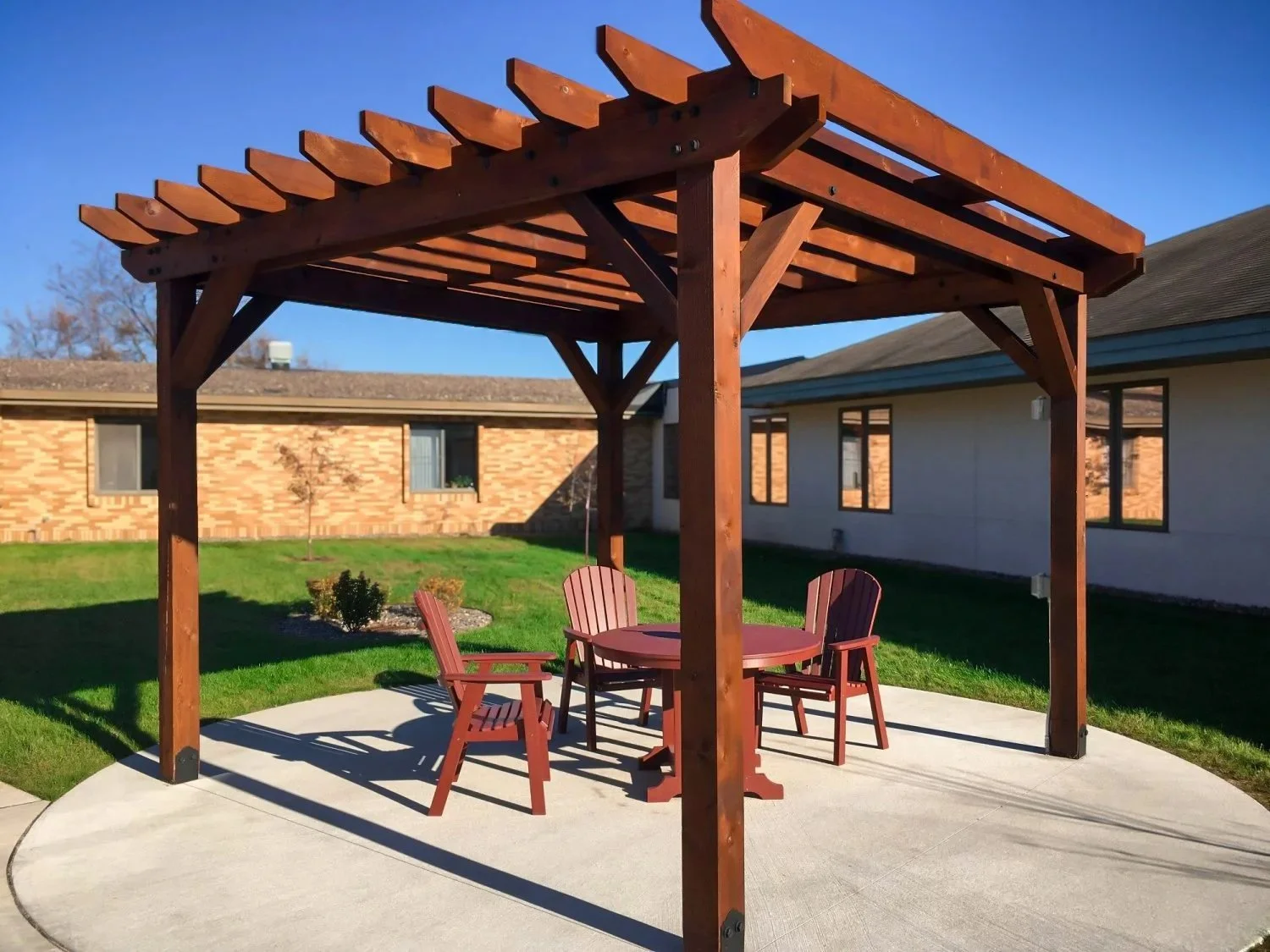 Wooden outdoor gazebo with round table and four chairs on a concrete patio in a backyard with green grass and nearby houses.