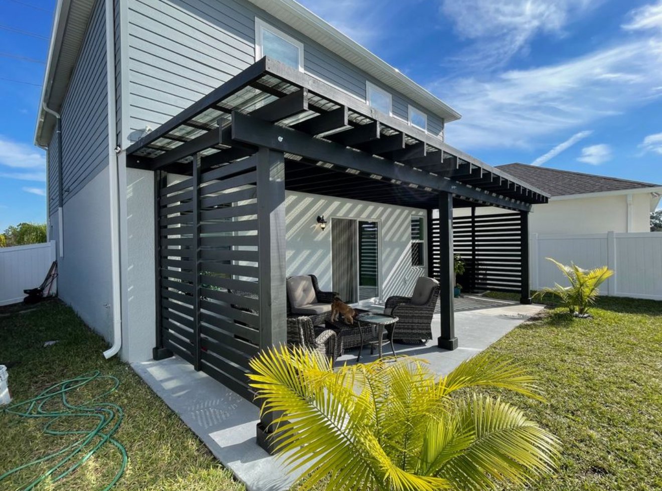 Backyard patio with black pergola, outdoor furniture, potted plants, and a house with light gray siding under a partly cloudy sky.