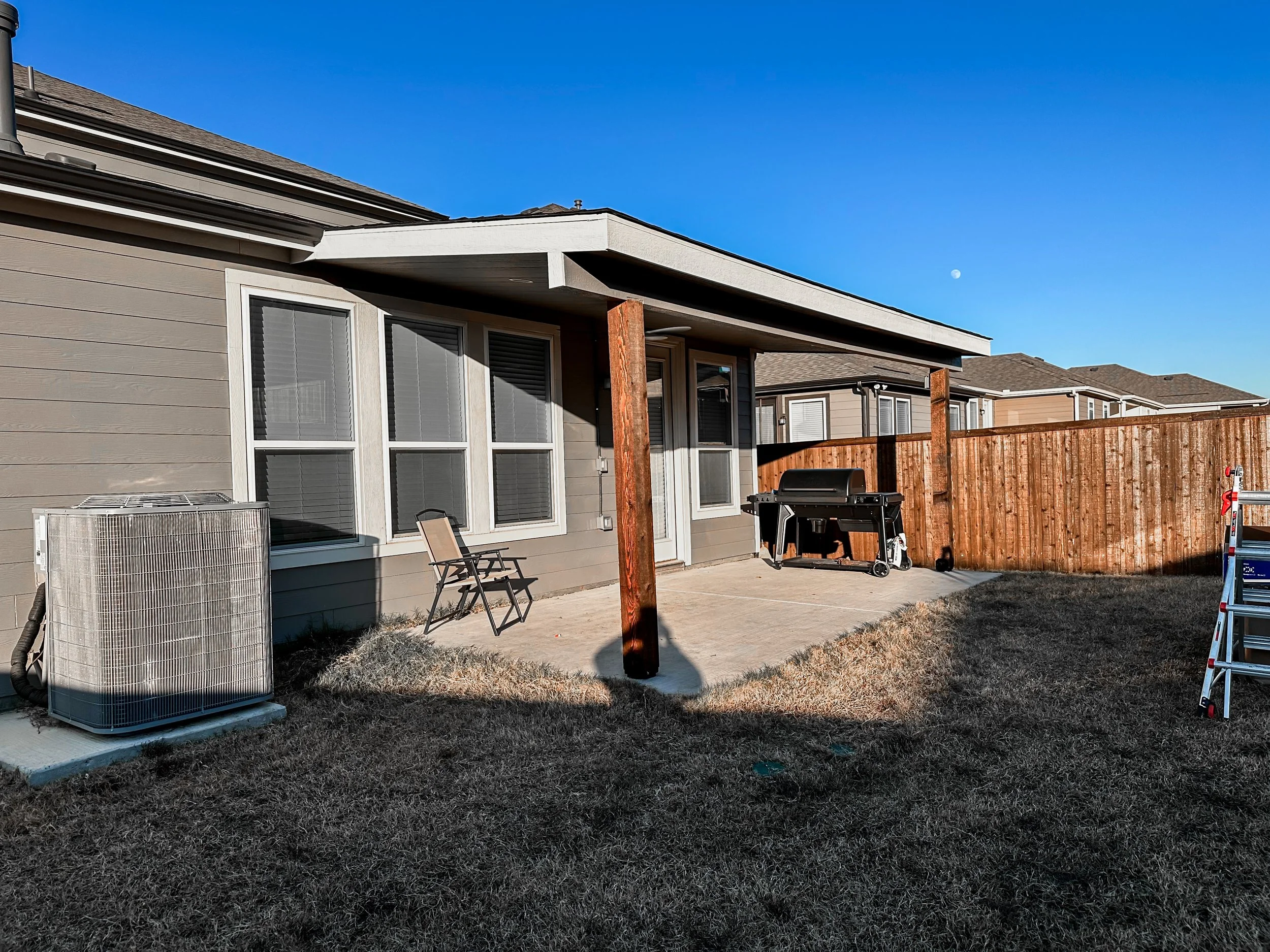 Backyard patio with beige house, large windows, air conditioning unit, barbecue grill, folding chairs, ladder, wooden fence, and dry grass under clear blue sky with visible moon.