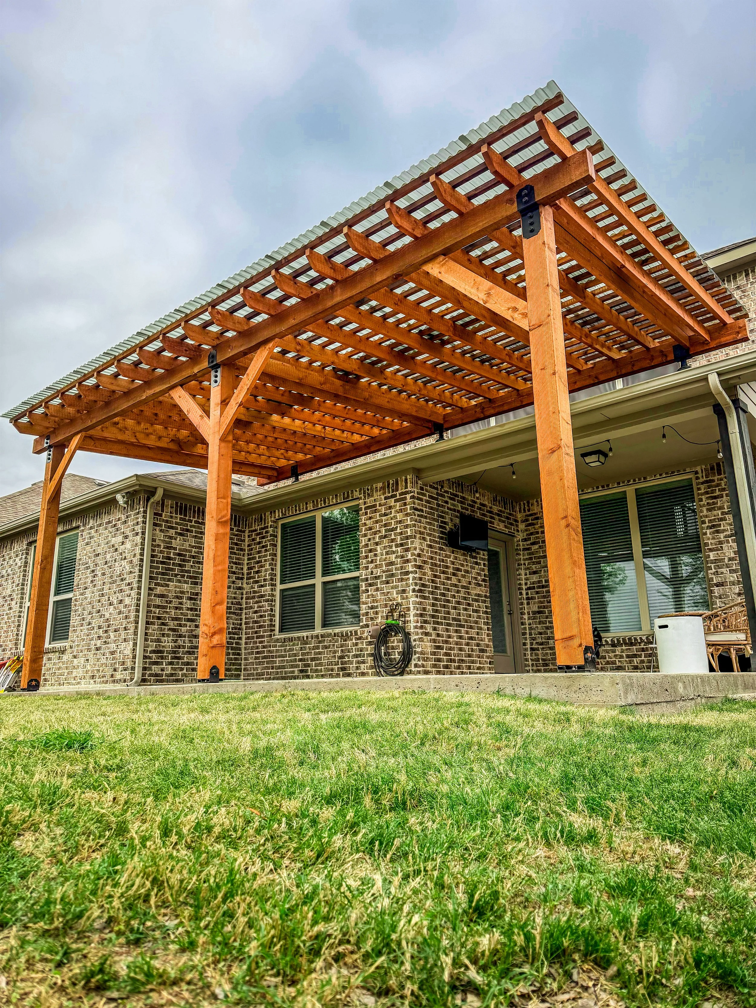 Backyard with brick house, glass windows, door, and a newly built wooden pergola with a corrugated roof over the patio area. Green grass in the foreground.