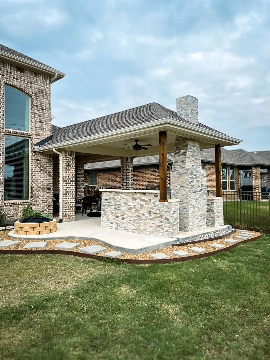 Backyard patio with stone and brick accents, decorated with pavers and gravel, with a covered area featuring a ceiling fan, barbecue grill, and outdoor seating, adjacent to a brick house.