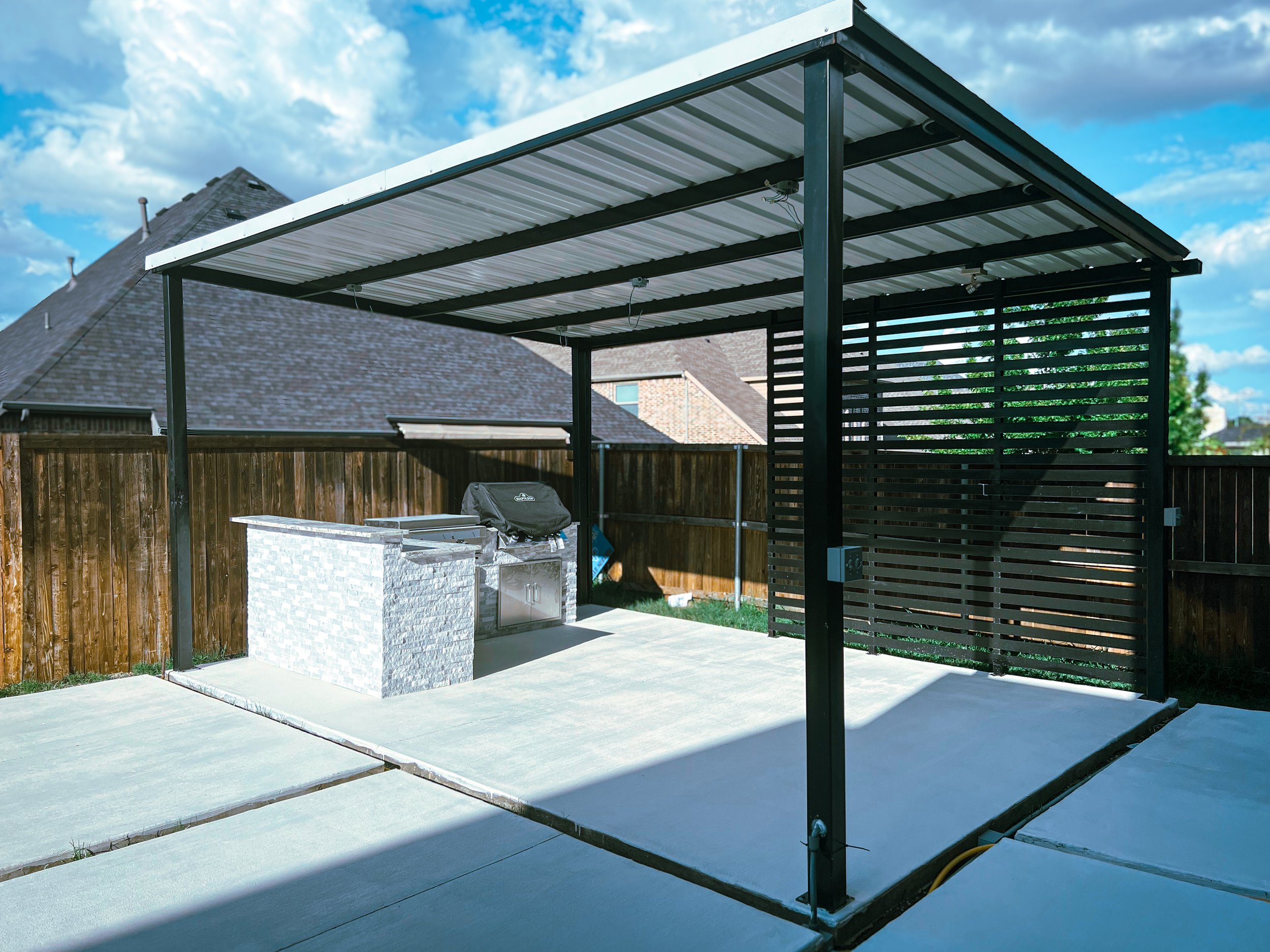 A backyard patio with a new metal and plastic pergola installed over a concrete slab, a barbecue grill covered with a black grill cover, and a wood fence in the background. Part of a house with a shingled roof is visible in the background.