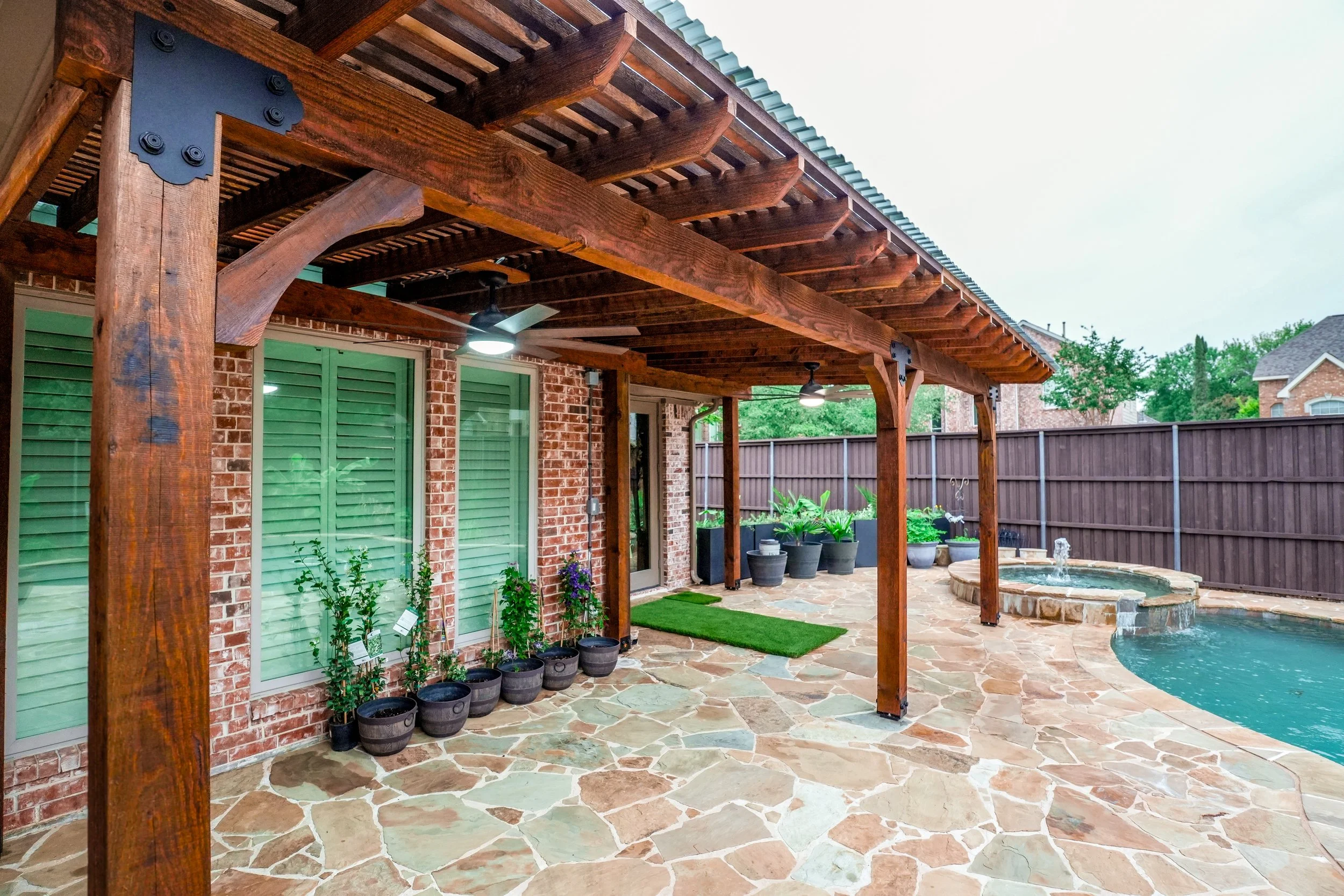 Backyard with patio, swimming pool, brick house with green shutters, wooden pergola, potted plants, and a water fountain.