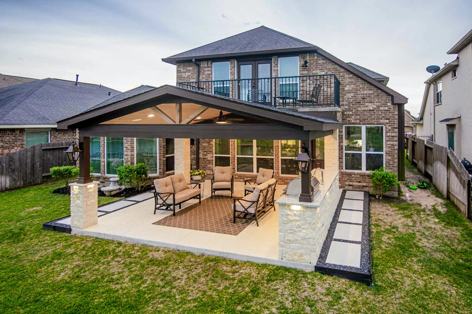 Backyard patio with seating area, covered by a pergola, with a house in the background featuring a upper balcony with chairs and a table.