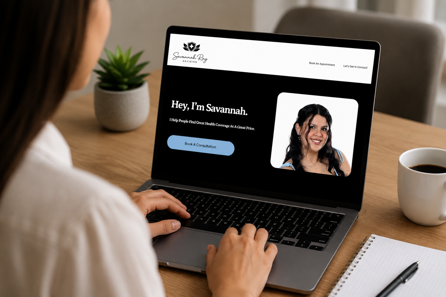 A woman sitting at a wooden desk browsing a health insurance website on her laptop. The website features a photo of Savannah Ray, a smiling woman with dark, wavy hair, and a button labeled 'Book A Consultation'. A potted plant, a cup of coffee, a notebook, and a pen are also on the desk.