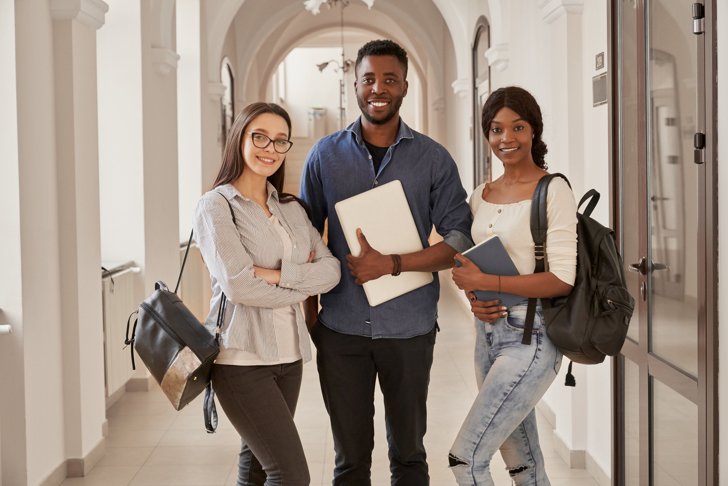 Three young adults standing in a hallway with lockers, smiling at the camera, holding notebooks and backpacks. Nigeria, ghana, africa