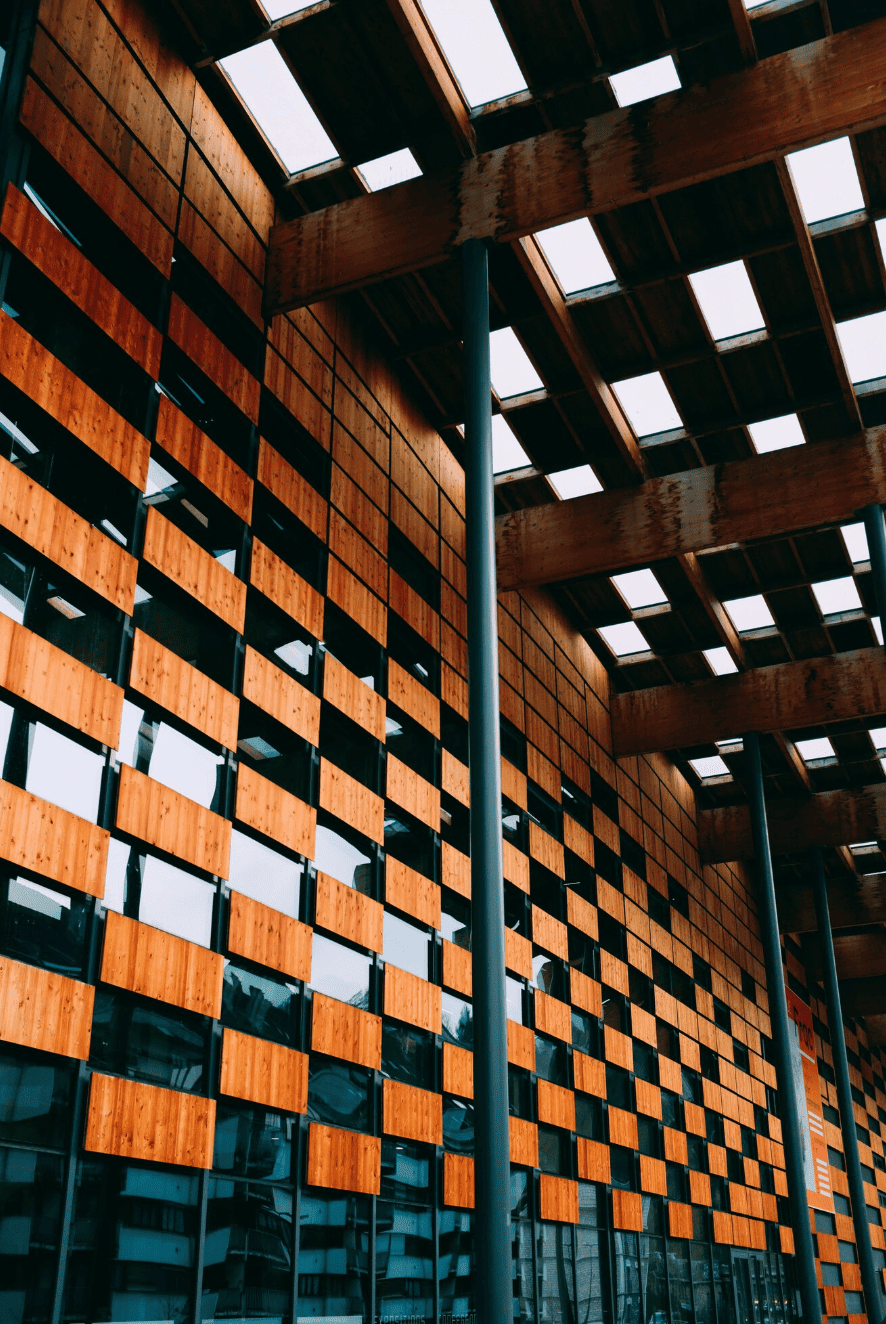 Modern building with a facade of wooden panels and large windows, topped with a wooden grid ceiling and skylights, supported by metal columns.