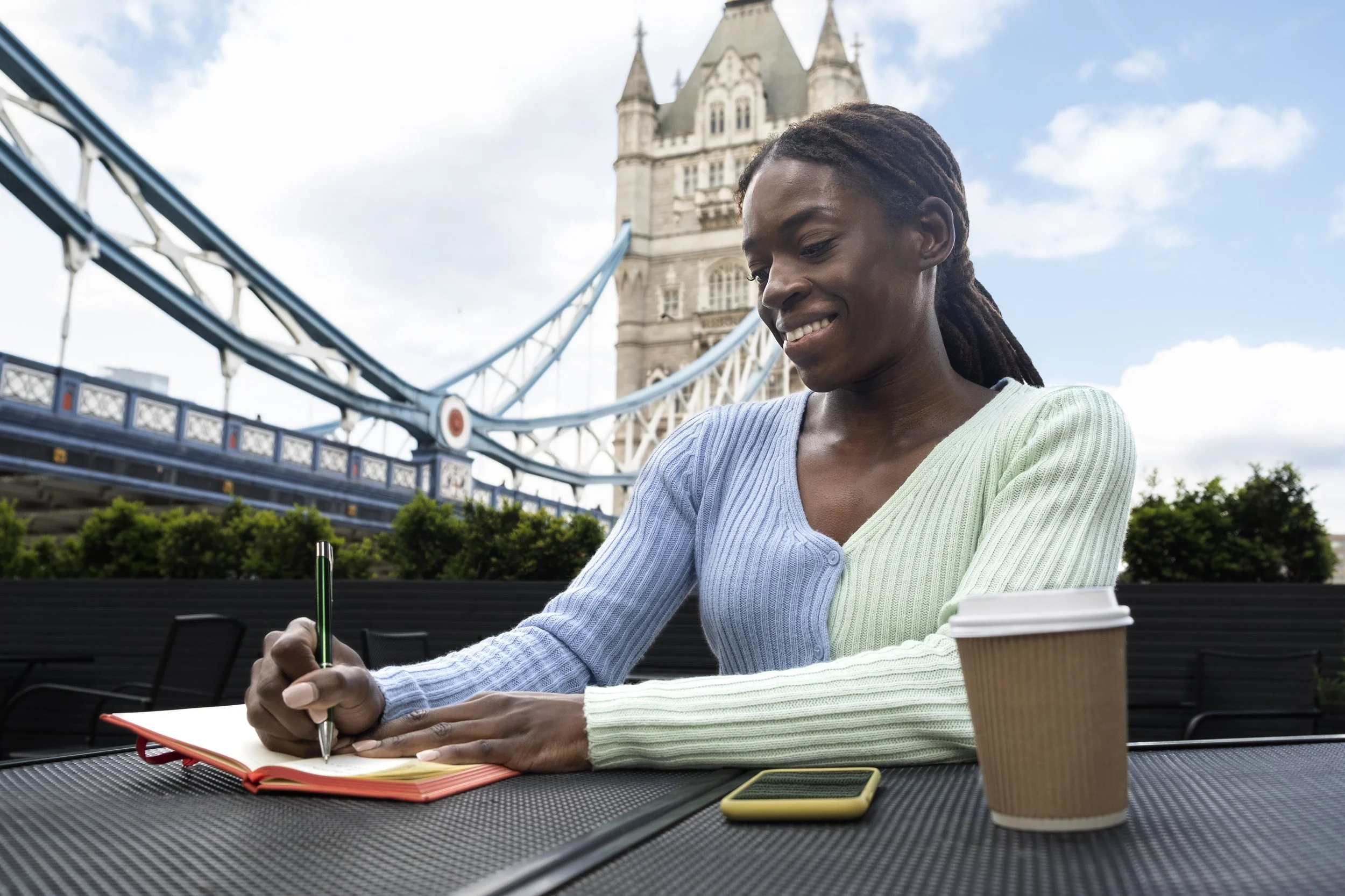 A woman with braided hair smiling as she writes in a notebook at an outdoor table, with Tower Bridge in London in the background on a partly cloudy day. She has a coffee cup and a smartphone on the table.