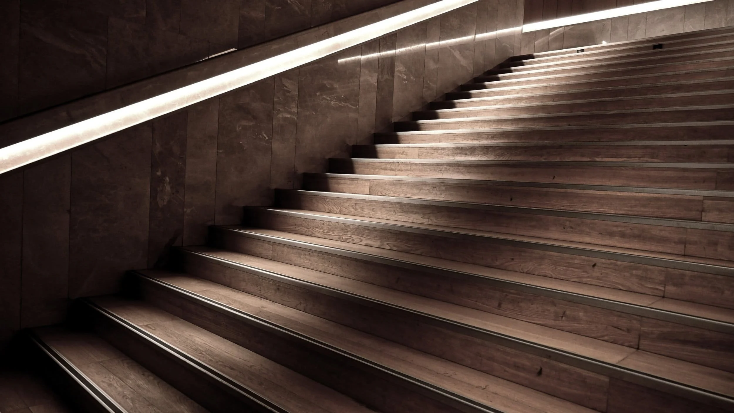 Wooden staircase with a modern handrail and dark walls, illuminated by overhead lighting.