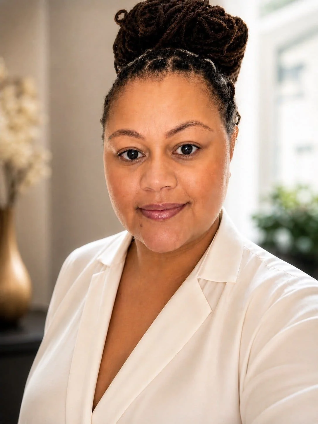 A woman with dark brown hair styled in thick dreadlocks tied up with a headband, wearing a cream-colored blazer, standing indoors near a window with blurred background. UK university admissions advisory Marsha Gayle