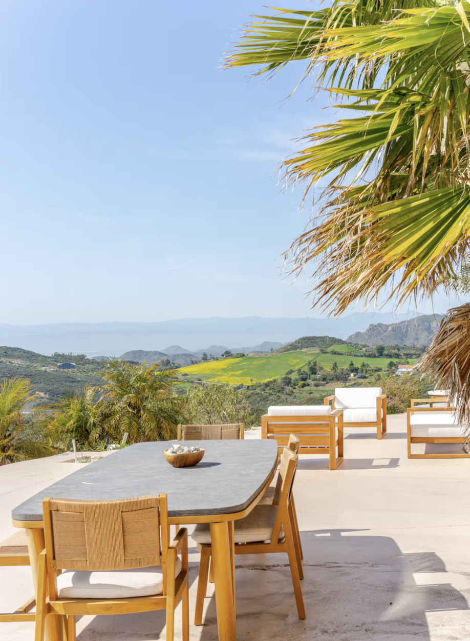 Outdoor patio with dining table and chairs, lounging chairs, overlooking a scenic landscape of hills and mountains, with palm trees and clear blue sky.