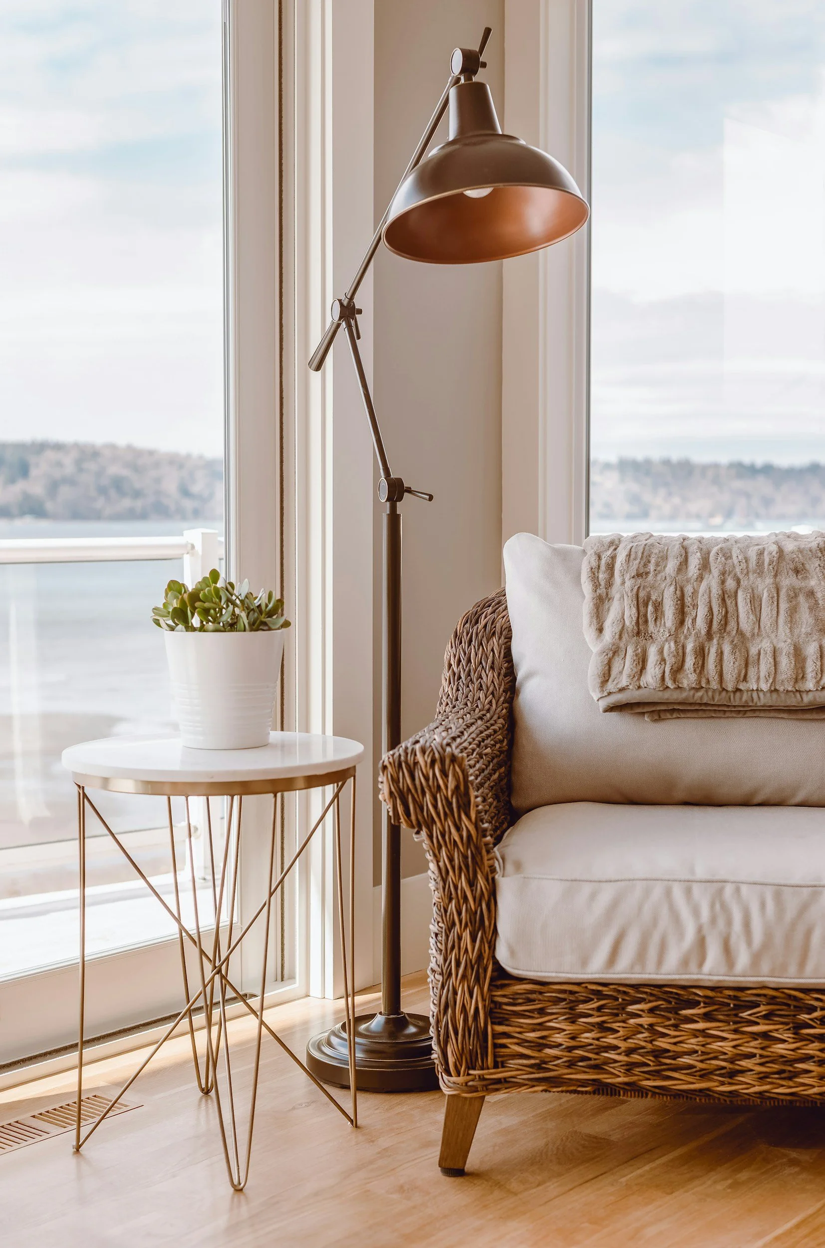 A cozy corner with a rattan armchair, a white throw pillow, a folded beige blanket, a small white side table with a potted succulent, a black adjustable floor lamp, and large glass doors with a view of a lake and distant trees.