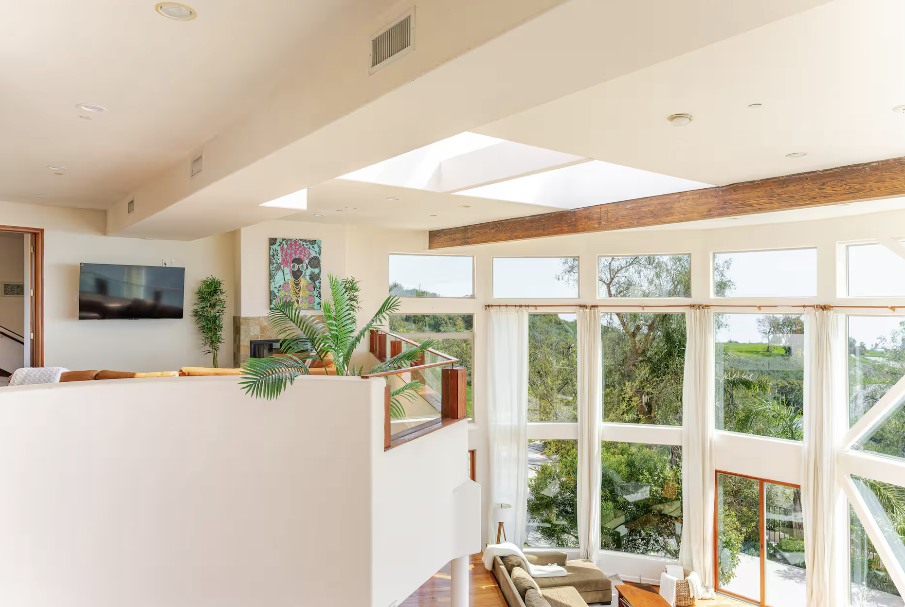 Bright living room with large windows, white curtains, and a view of trees outside. Features include a brown sectional sofa, a flat-screen TV, potted plants, colorful artwork, and wooden accents on the ceiling and handrails.