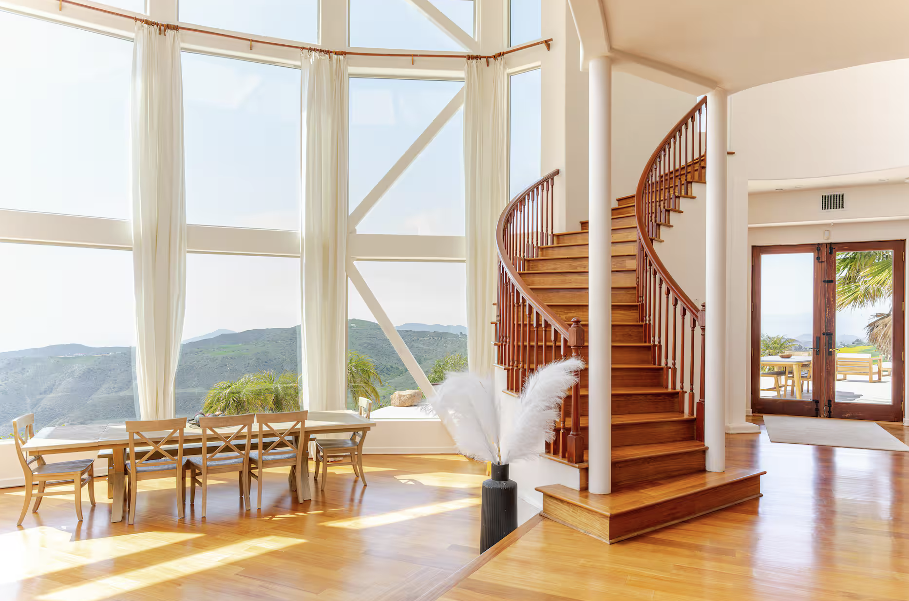 Interior view of a spacious home with large windows showing a scenic mountain view, a wooden spiral staircase, a dining table with chairs, and a sliding glass door leading to an outdoor patio with seating.