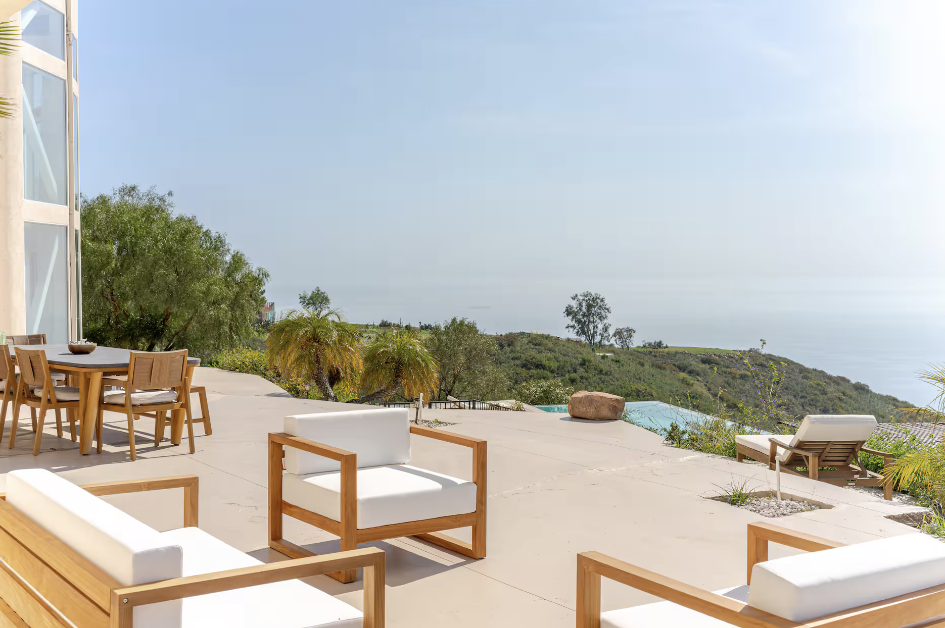 Modern outdoor patio with white and wooden seating, surrounded by palm trees and overlooking a hillside and ocean in the background.