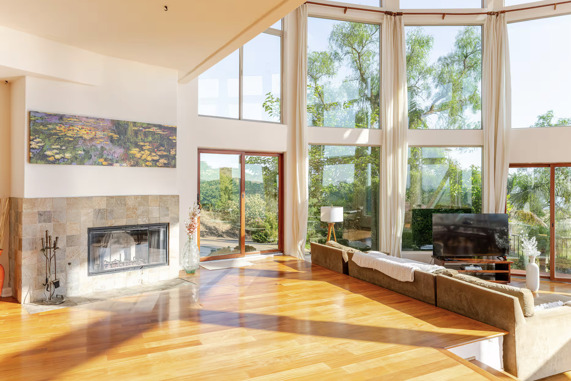 Bright living room with large windows, white curtains, a fireplace with stone surround, a flat-screen TV, a brown sofa, a coffee table, and decorative plants, with sunlight streaming in and views of green trees outside.