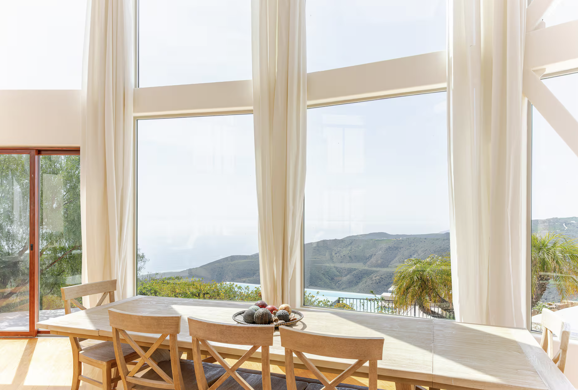 Dining room with large windows showing mountain landscape, beige curtains, a wooden dining table, and chairs, with a decorative bowl of balls on the table.