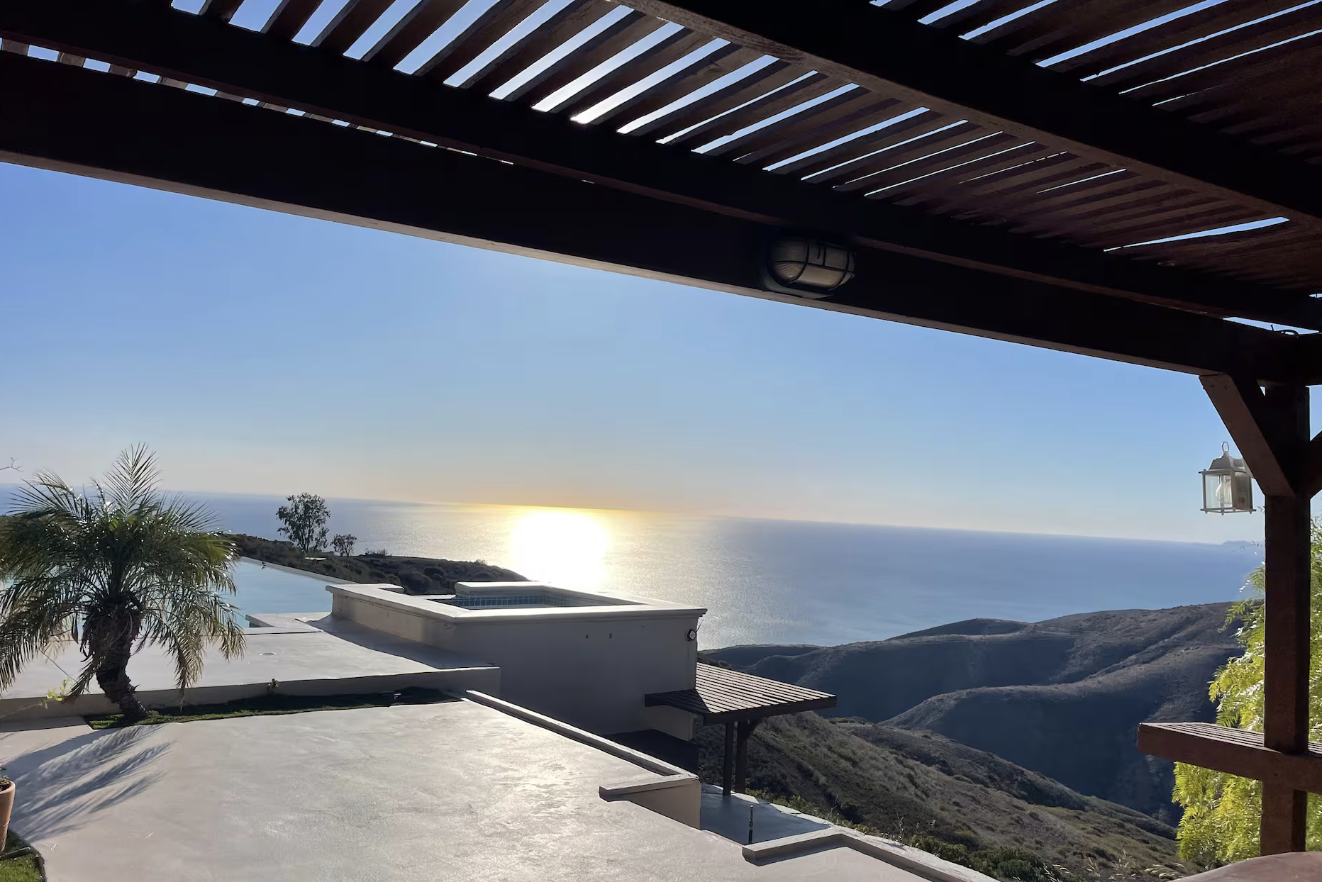 View of the ocean at sunset from a balcony with a pergola, palm tree, and hillside landscape.