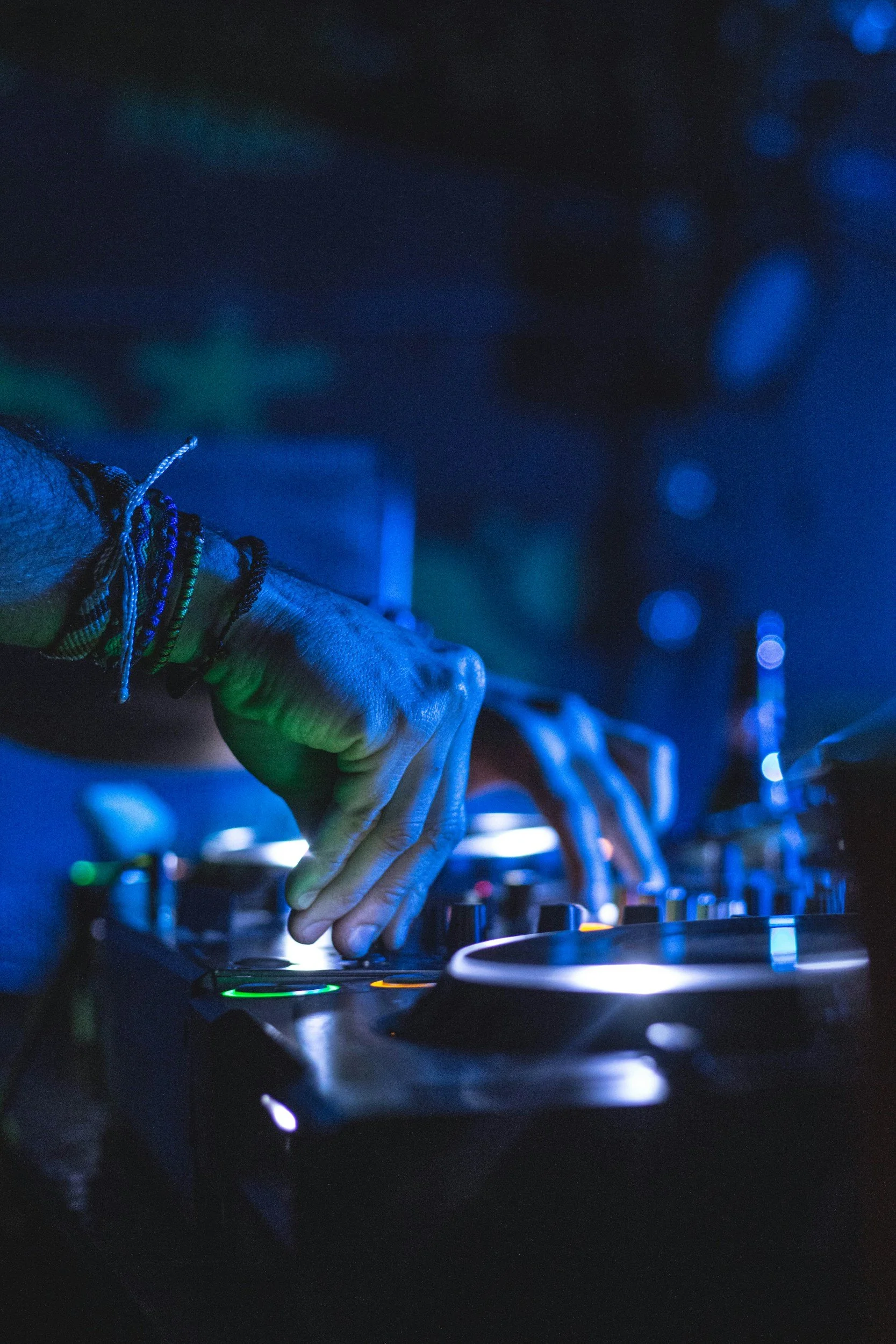 A DJ's hand adjusting a mixer knob with colorful bracelets on the wrist, in a dark club setting with blue lighting.
