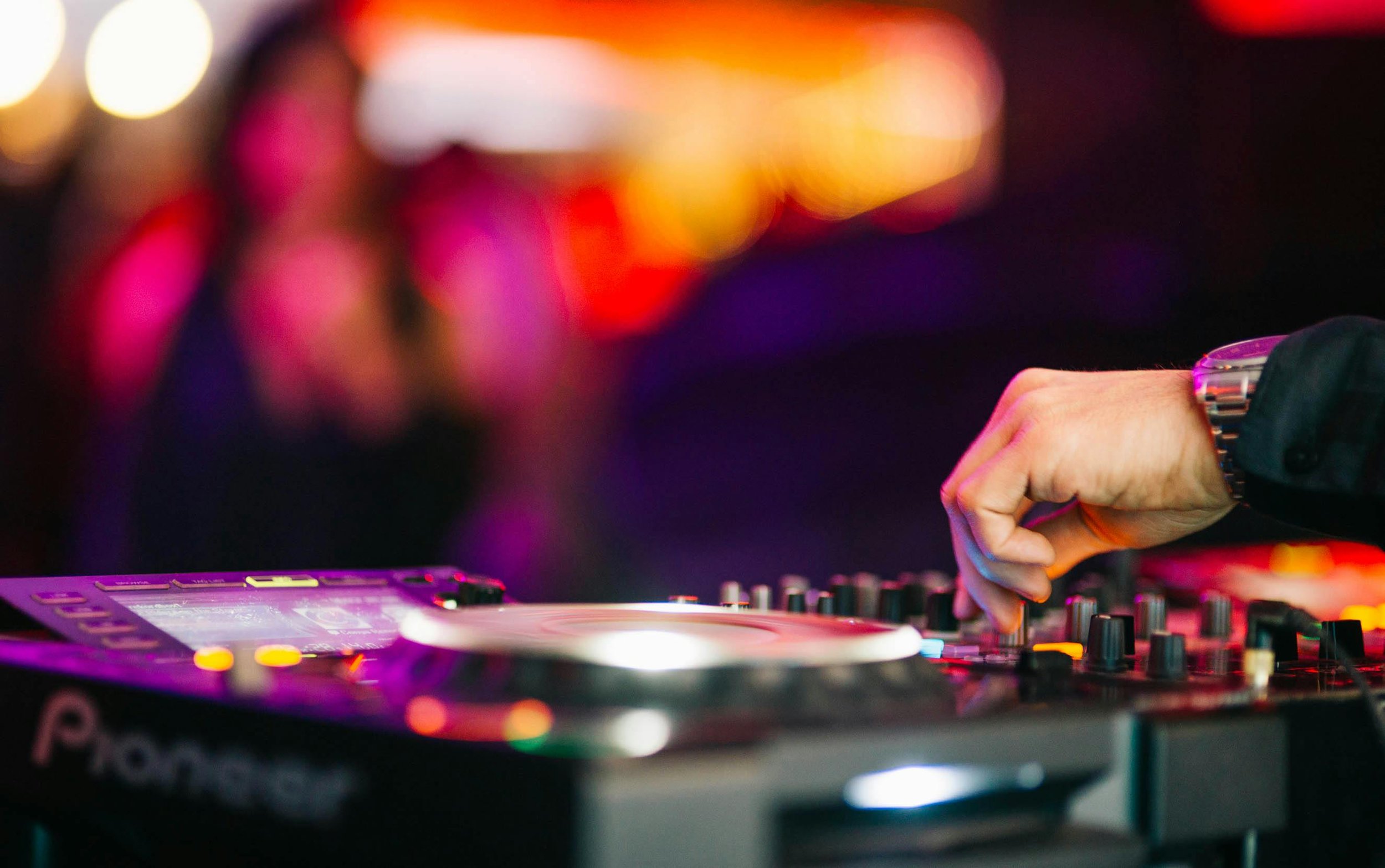 A DJ's hand adjusting knobs on a DJ mixer at a nightclub, with colorful blurred lights in the background.