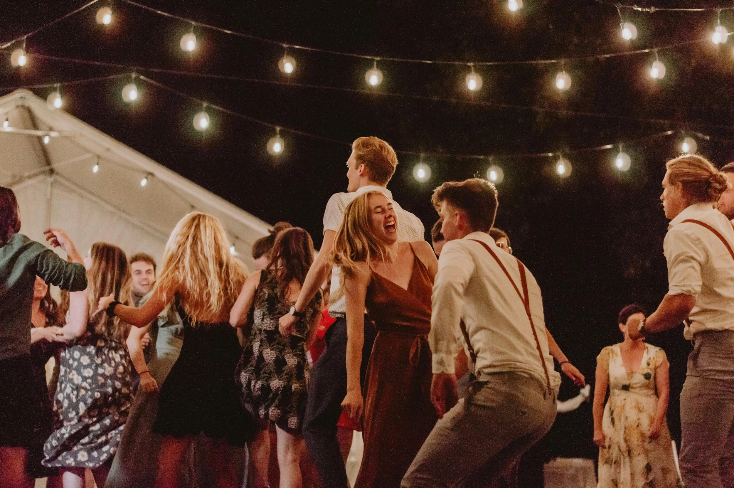 People dancing and enjoying themselves at an outdoor party under string lights.