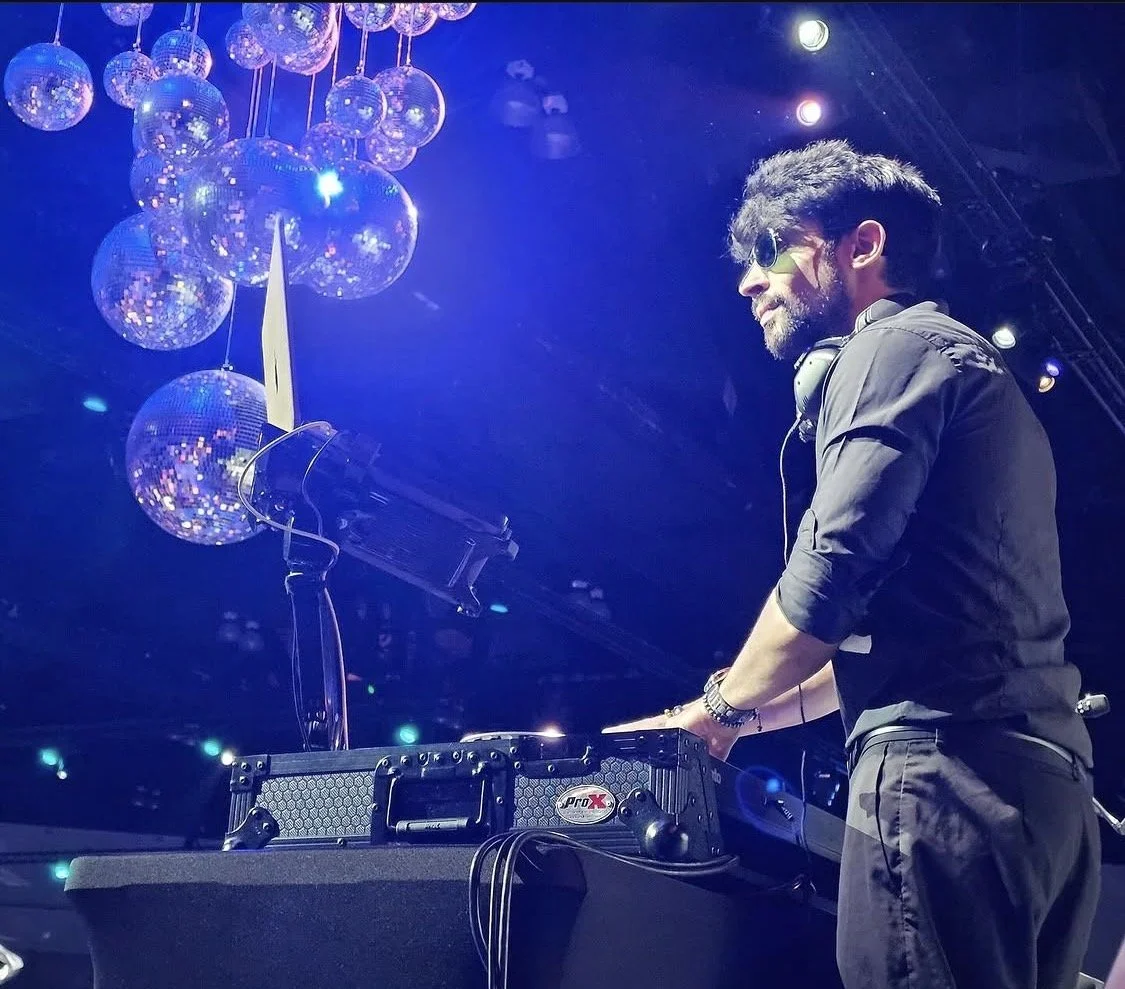 DJ Burno with headphones playing music at a dance party, illuminated by blue and purple lights with hanging disco balls in the background.