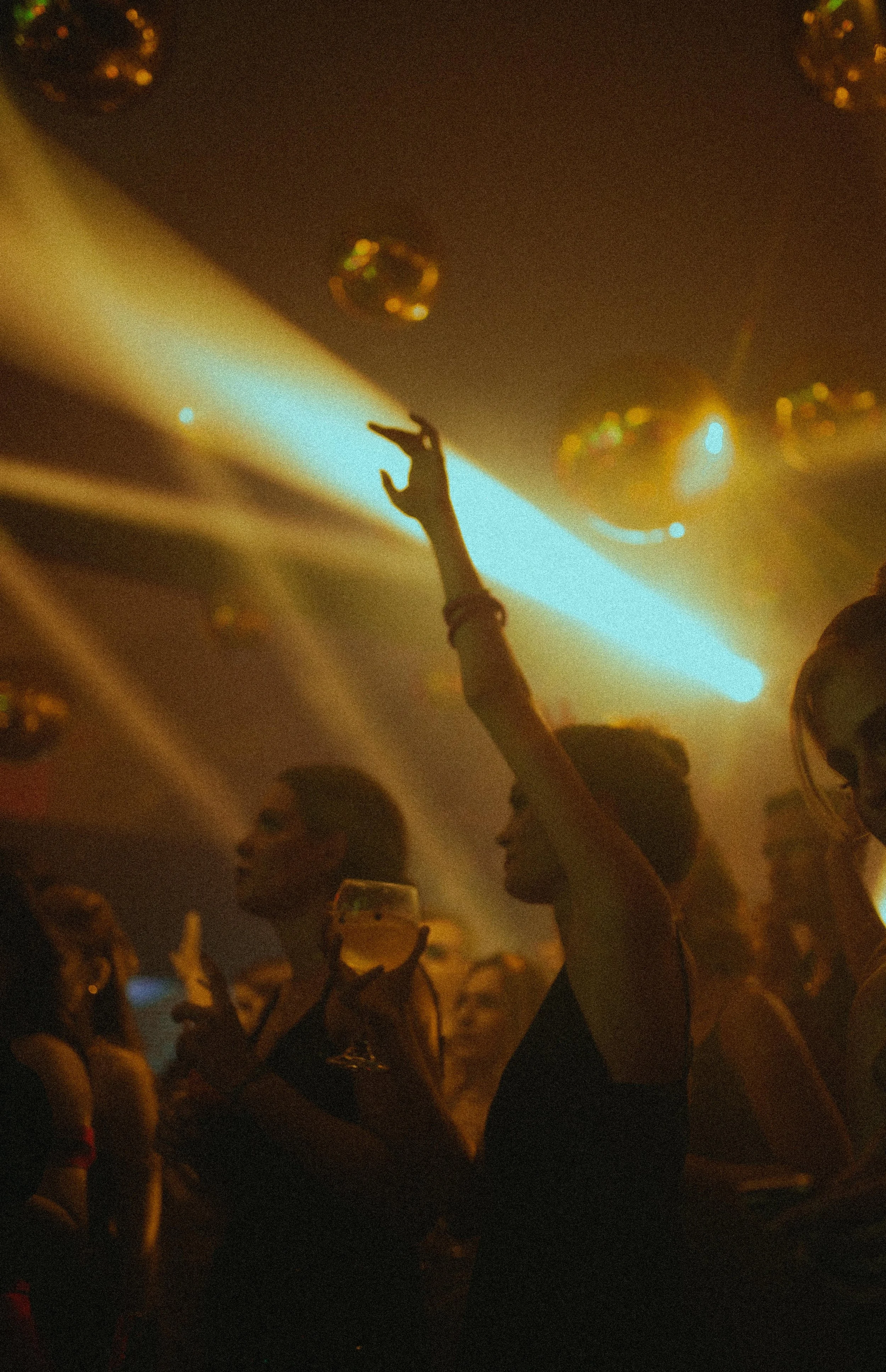 Crowd of people at a large event with a person in the foreground reaching up towards the stage, illuminated by stage lights and spotlights