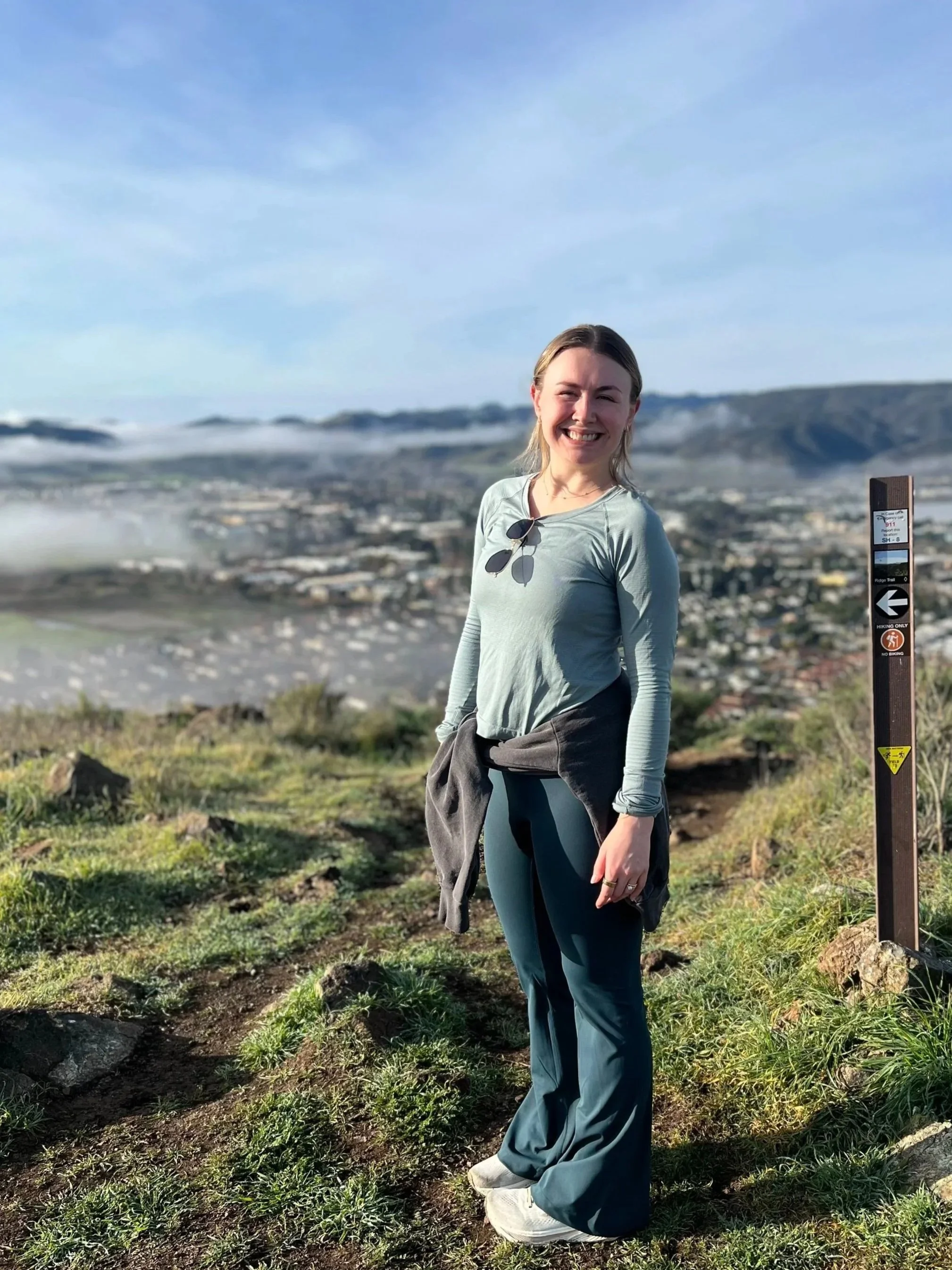 Smiling young woman in outdoor hiking attire standing on a trail with a scenic city and mountain view in the background. There is a trail marker to her right.