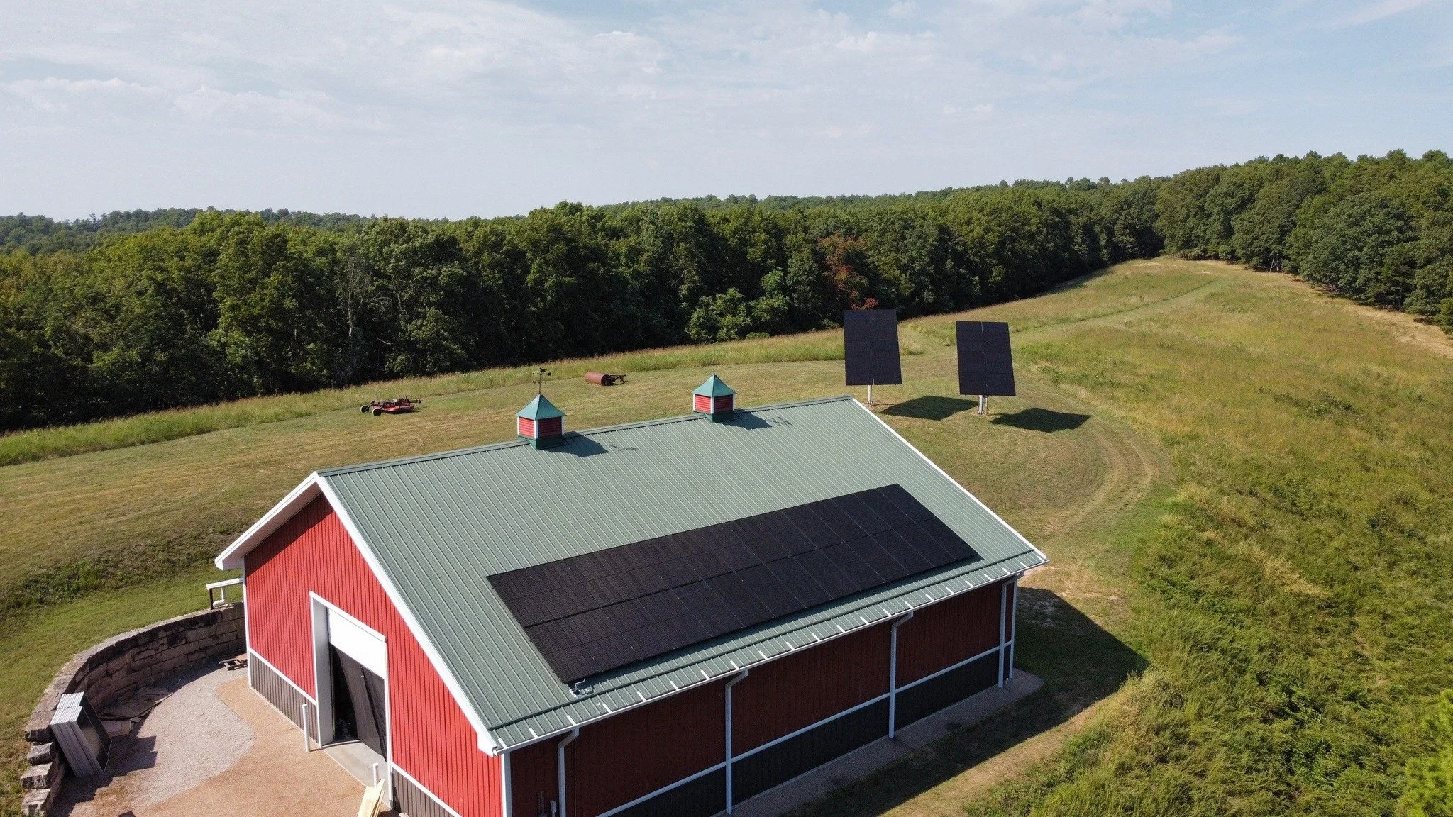 A red barn with a green metal roof in a rural area, solar panels are installed on the roof, and solar panels are also positioned on the ground nearby. The barn is surrounded by grass with a forest in the background, under a partly cloudy sky.