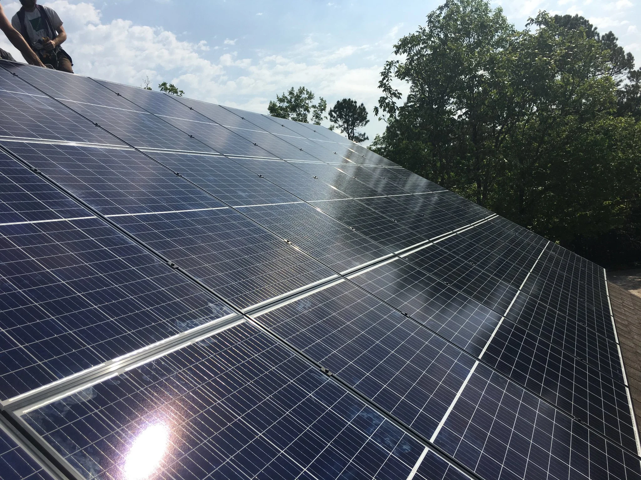 Photograph of solar panels installed on a roof with trees and a partly cloudy sky in the background.