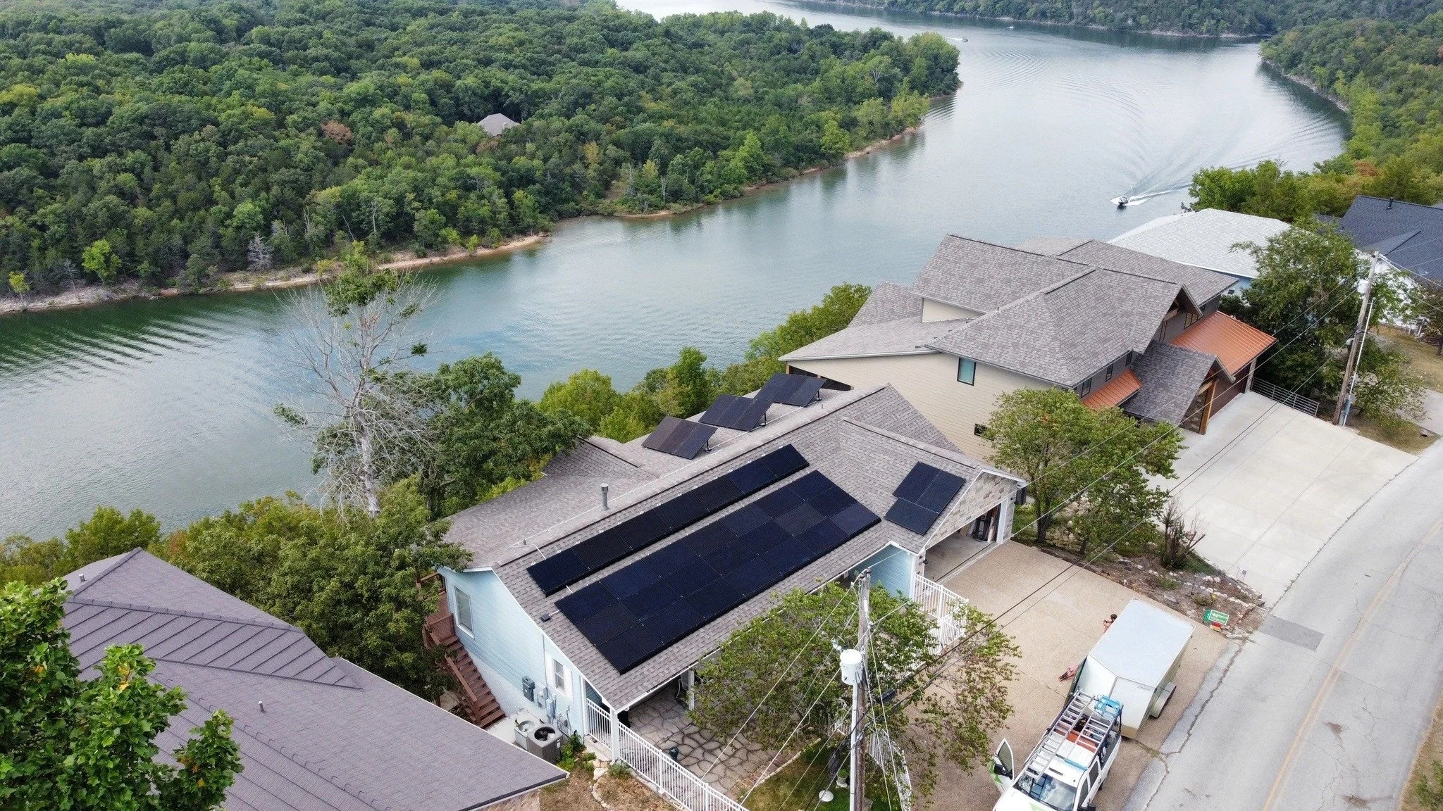 Aerial view of houses with solar panels on the roofs, next to a river with dense green trees on the opposite bank.