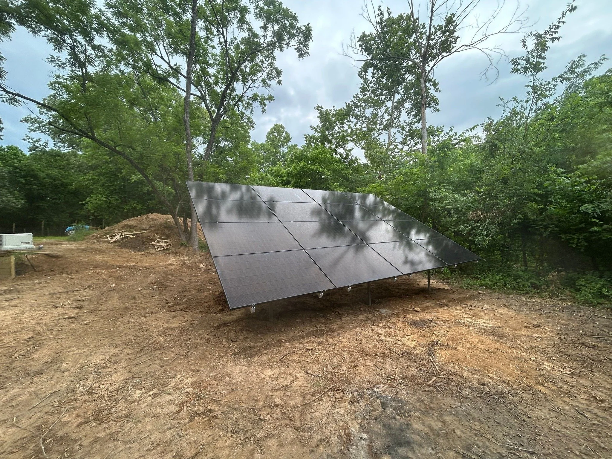 Solar panels installed on the ground in a cleared outdoor area surrounded by trees and cloudy sky.