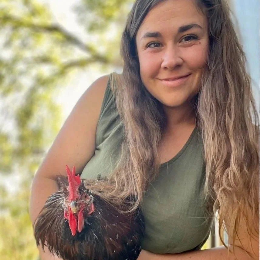 A woman with long, wavy brown hair smiling and holding a rooster outdoors.