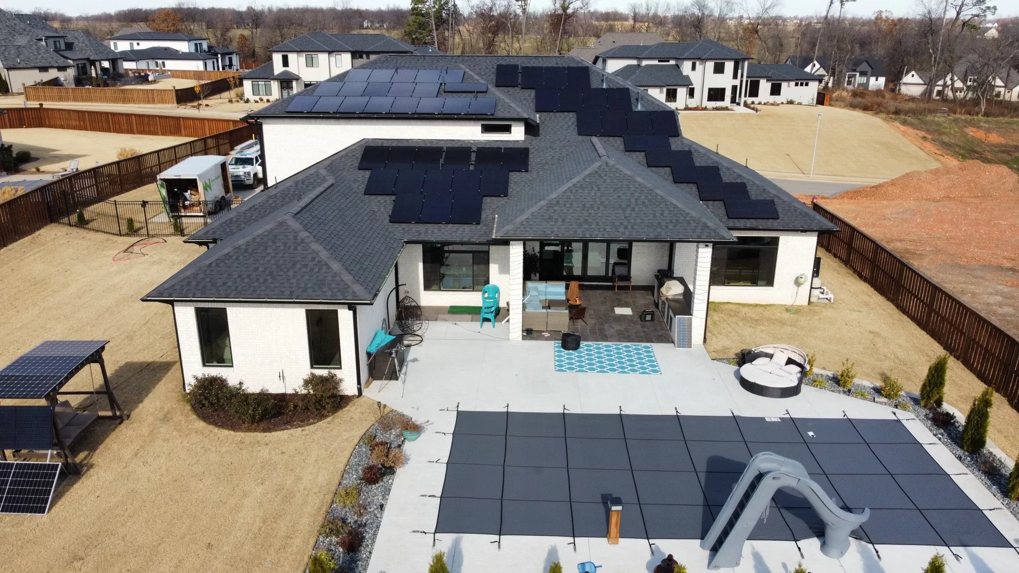 Aerial view of a modern house with a roof covered in black solar panels, backyard patio with outdoor furniture, and neighboring houses with similar roofing in a suburban neighborhood.