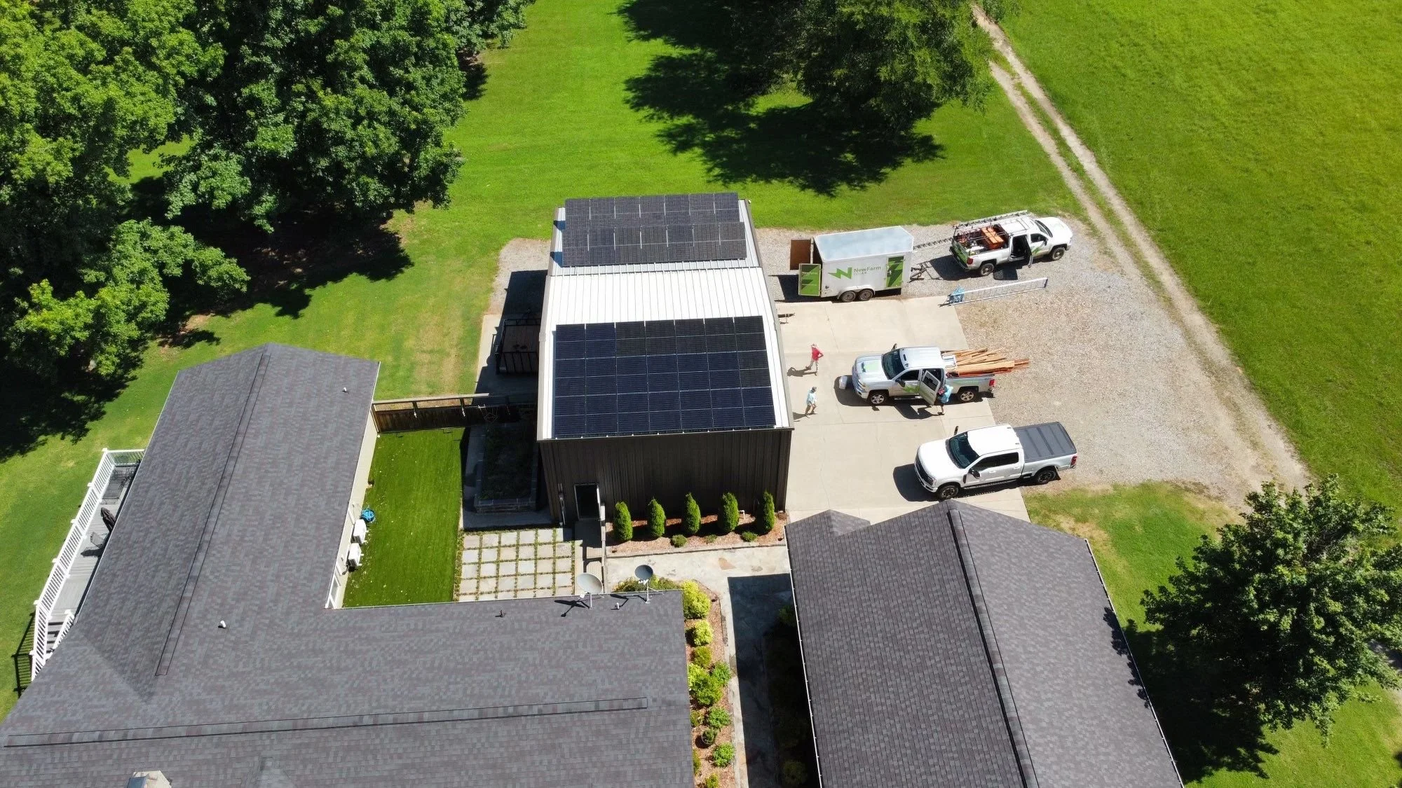 Aerial view of a house with solar panels on the roof, three pickup trucks in the driveway, and green lawn with trees surrounding the property.
