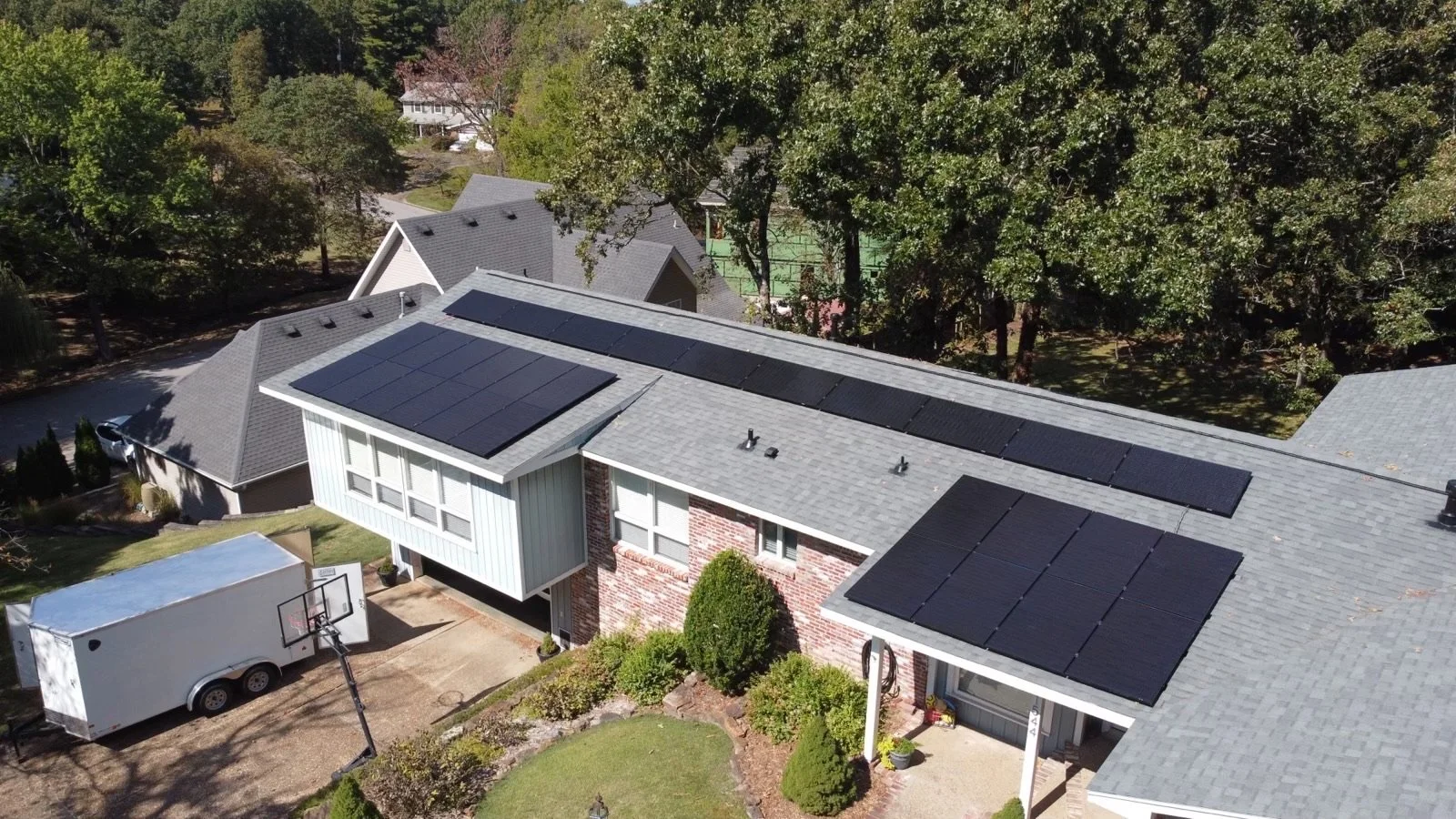 Aerial view of a house with solar panel installation on the roof, surrounded by trees and neighboring houses.