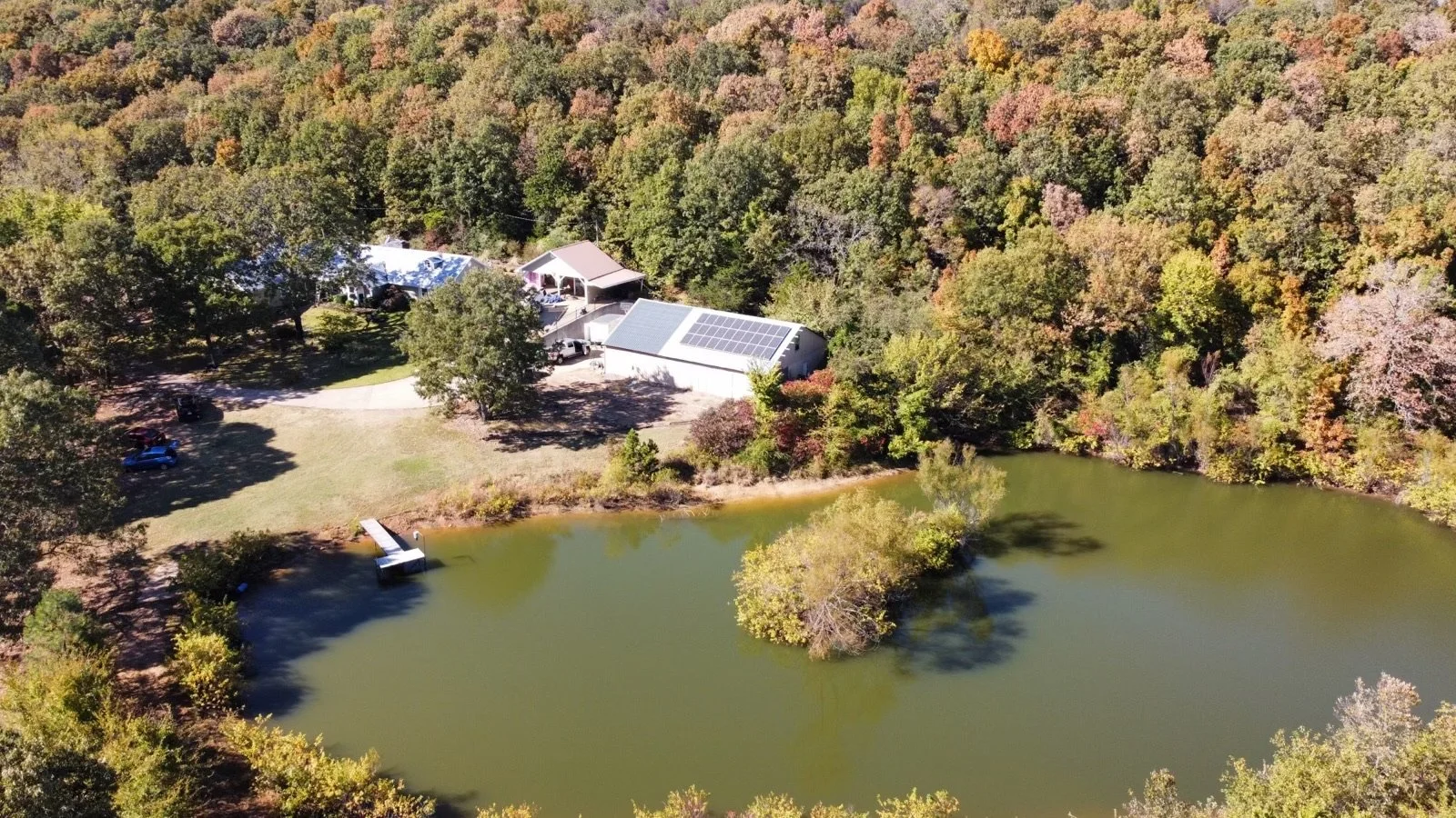 Aerial view of a lakeside property with several buildings, a dock, and surrounded by trees with fall foliage.