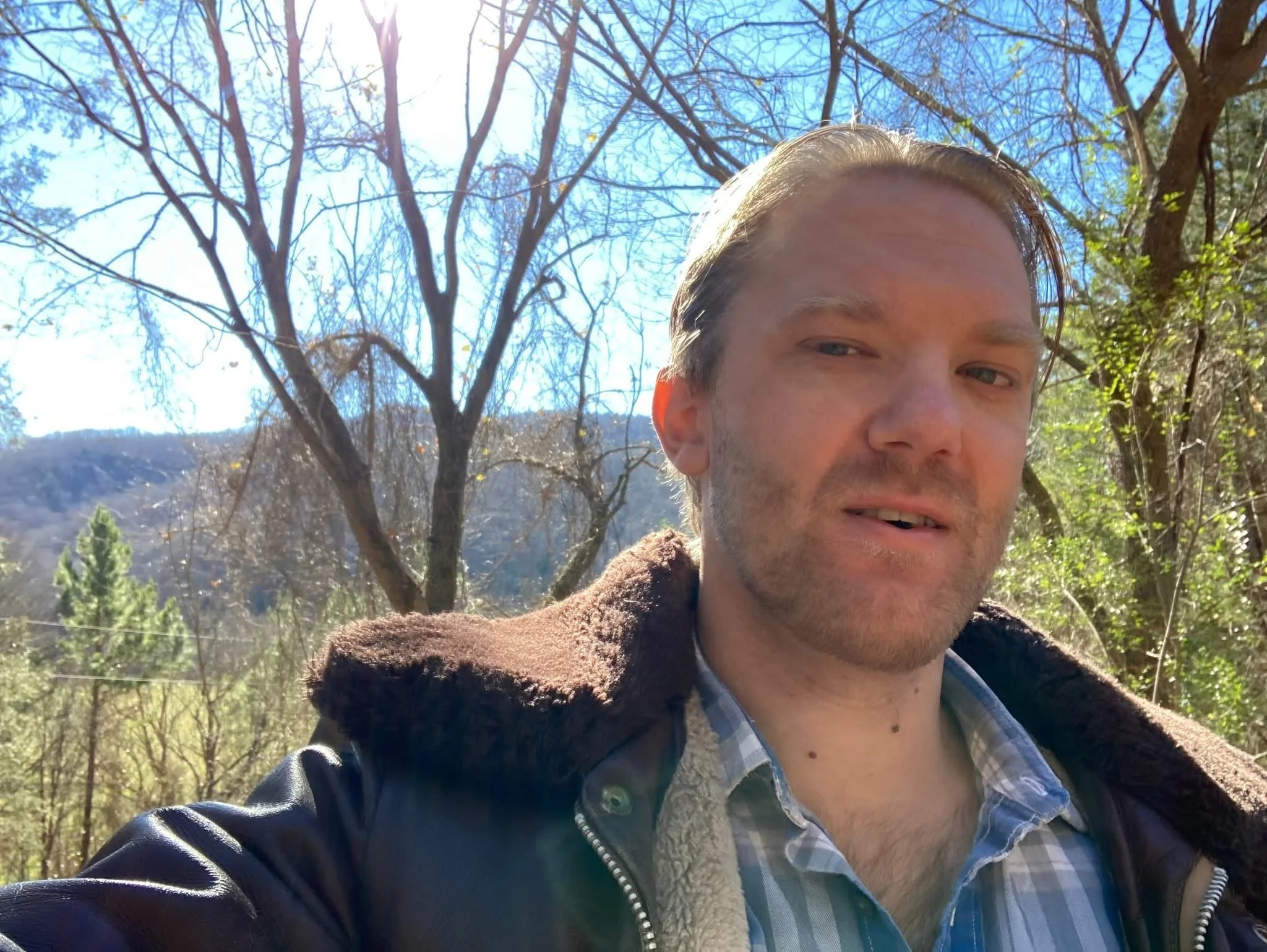 Close-up of a young man with light brown hair and facial hair, wearing a checkered shirt under a brown jacket, outdoors on a sunny day with leafless trees and mountains in the background.