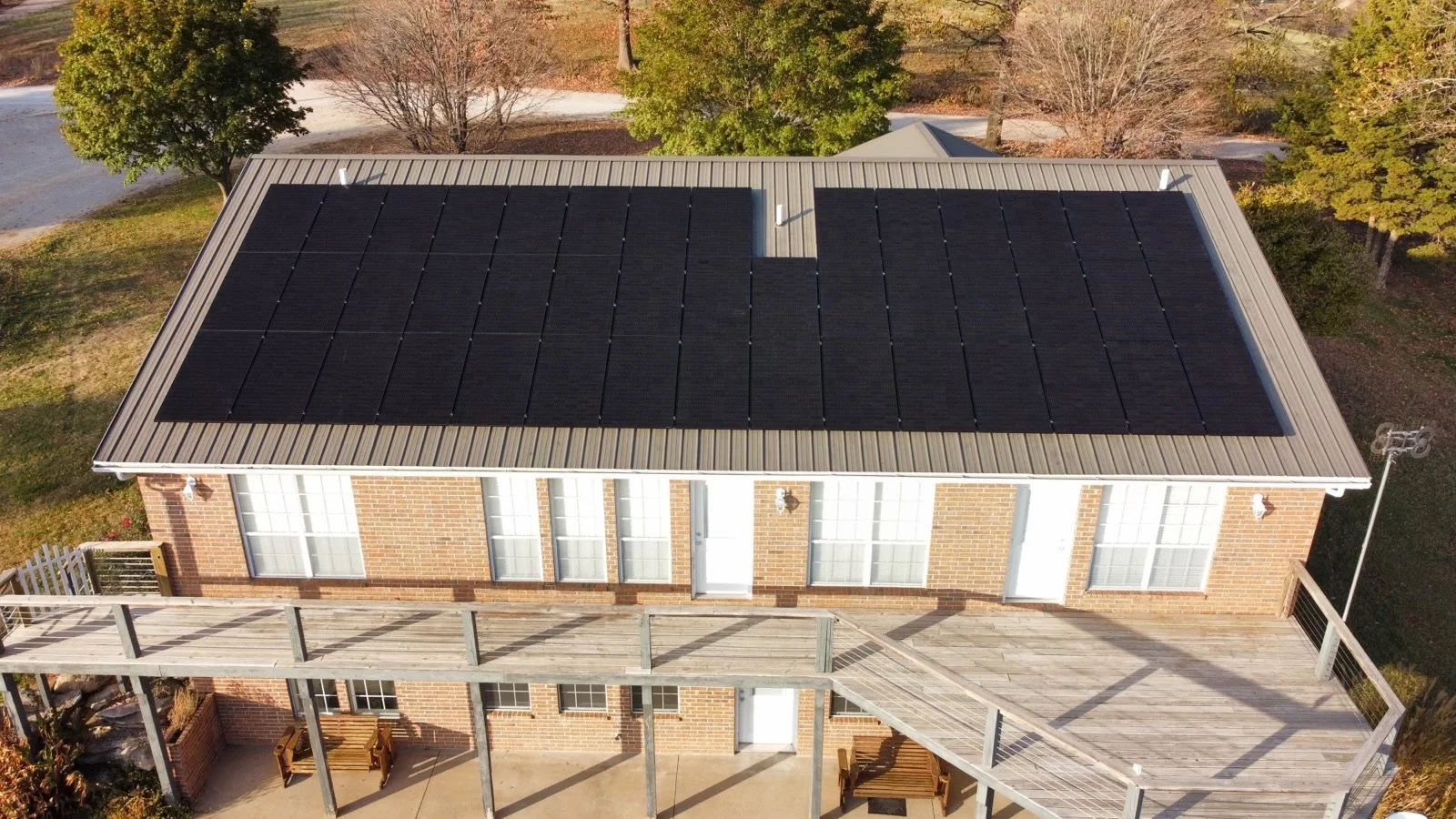 Aerial view of a brick house with a metal roof, featuring multiple solar panels installed on one side, and a large wooden balcony with benches and outdoor seating.
