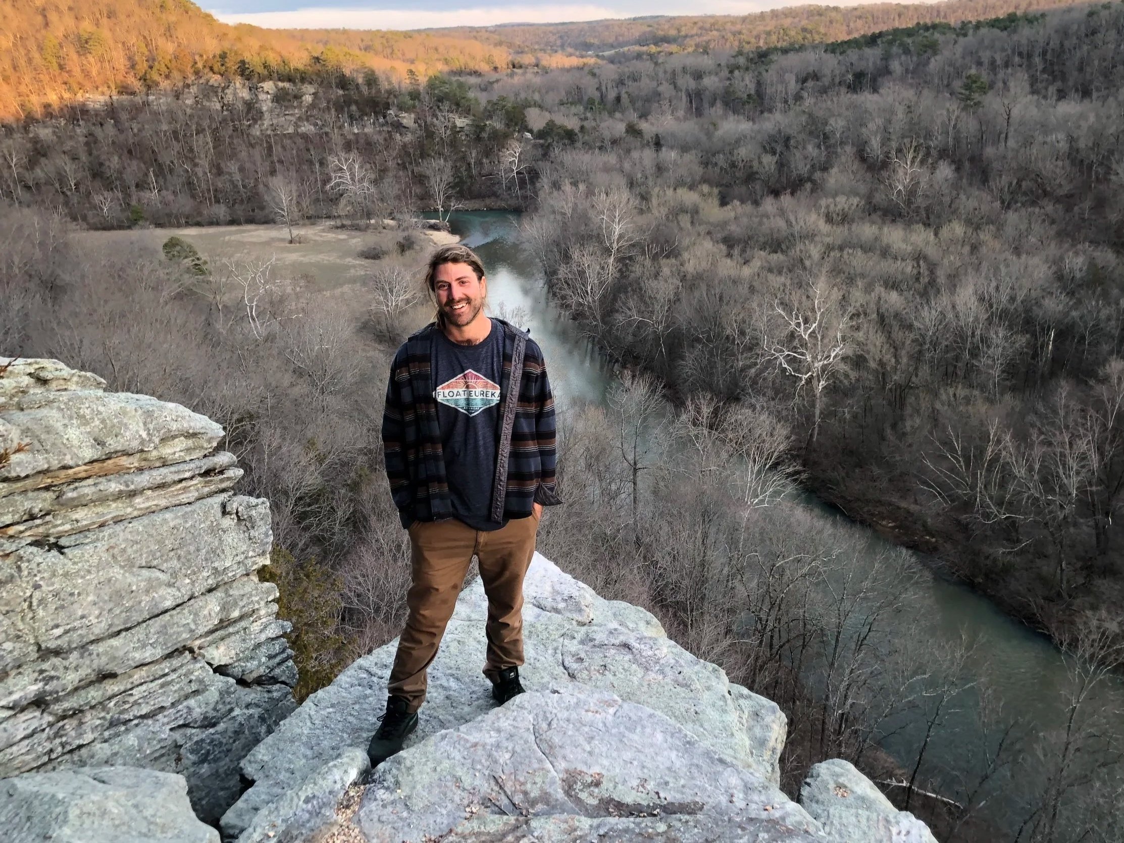 A man with long hair and a beard smiling, standing on rocky terrain near a river and surrounded by leafless trees, with hills and a river in the background during sunset.
