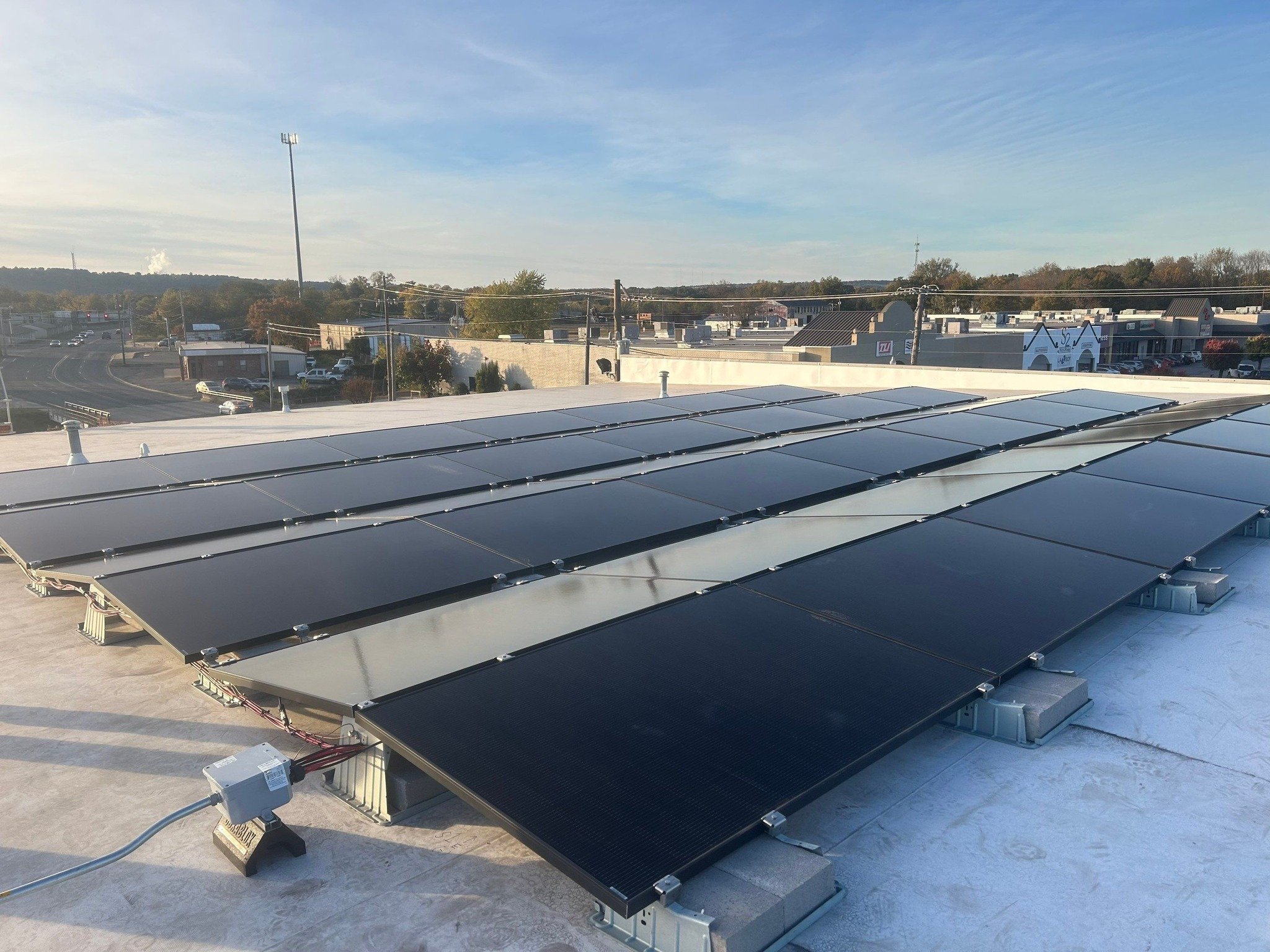 Multiple black solar panels installed on a rooftop with a view of a commercial area and distant trees under a blue sky.