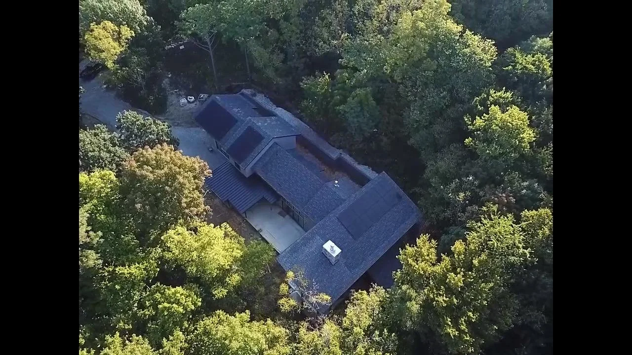 A large house surrounded by dense green trees, with a dark roof, on a wooded lot.